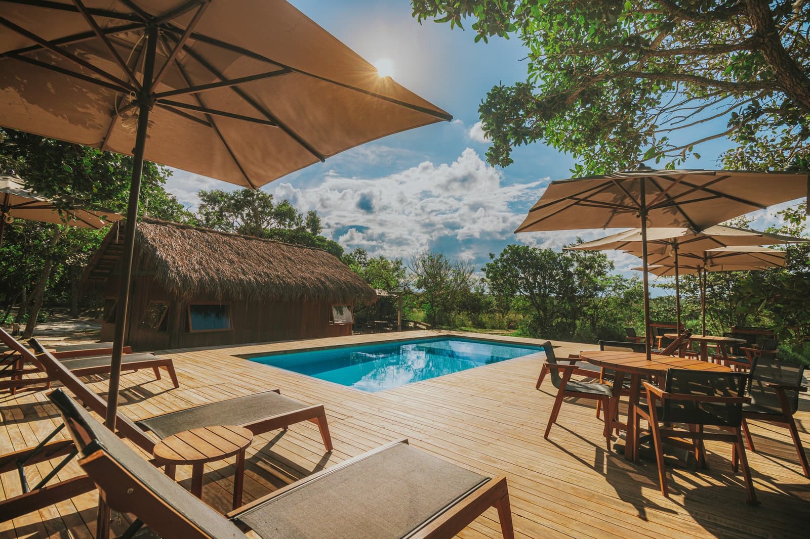 Um mergulho, uma sombra, e o cerrado logo ali do outro lado. Deck de madeira ensolarado com piscina na Pousada Trijunção, rodeado pela vegetação do Cerrado, espreguiçadeiras e sombrinhas sob um céu azul