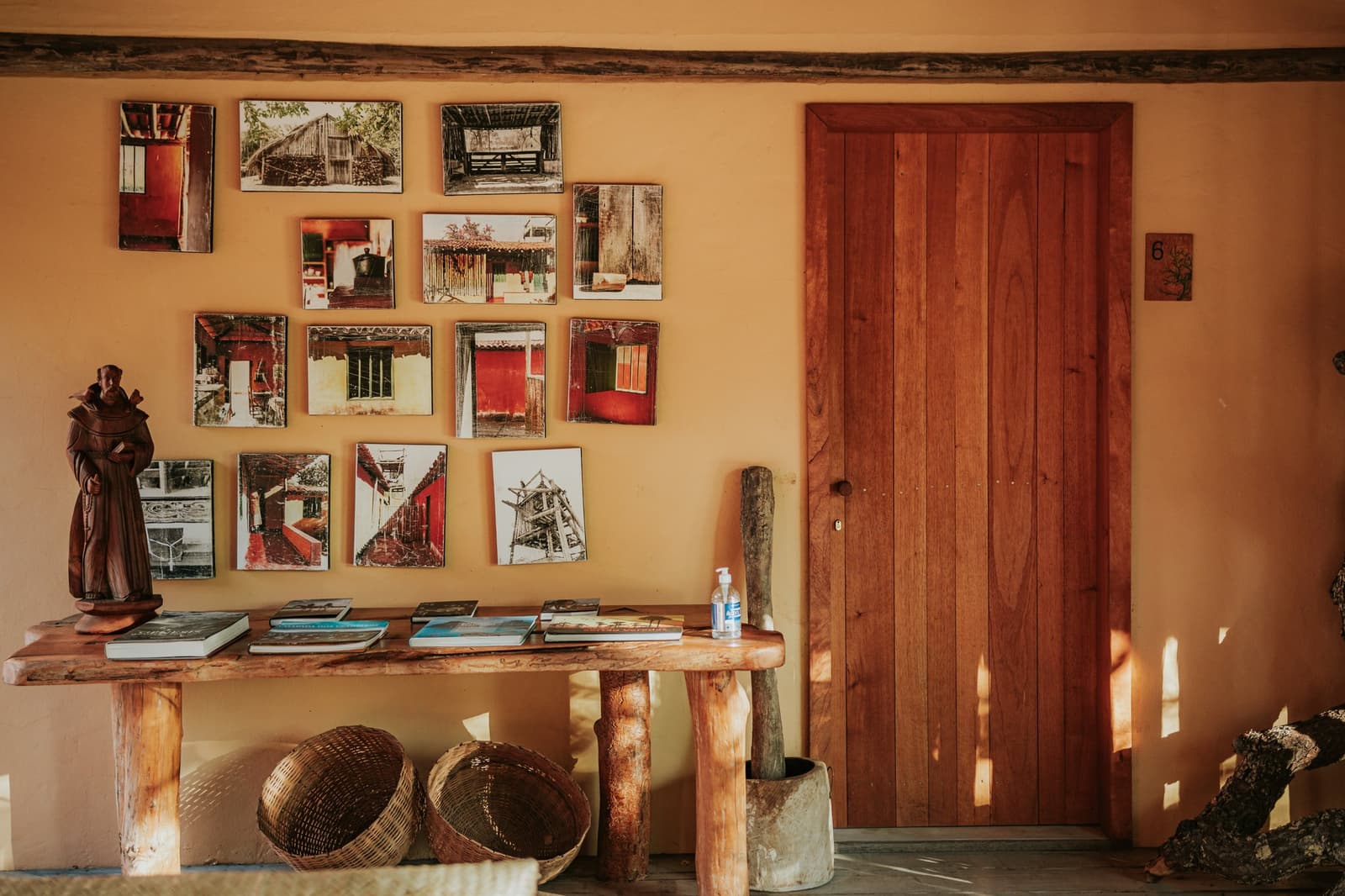 Cada foto nessa parede tem uma história que o pessoal da pousada conta com prazer. Parede em tom ocre da Pousada Trijunção decorada com uma galeria de fotografias retratando arquitetura tradicional do Cerrado, ao lado de uma mesa de madeira artesanal com livros, uma escultura de monge e cestos trançados