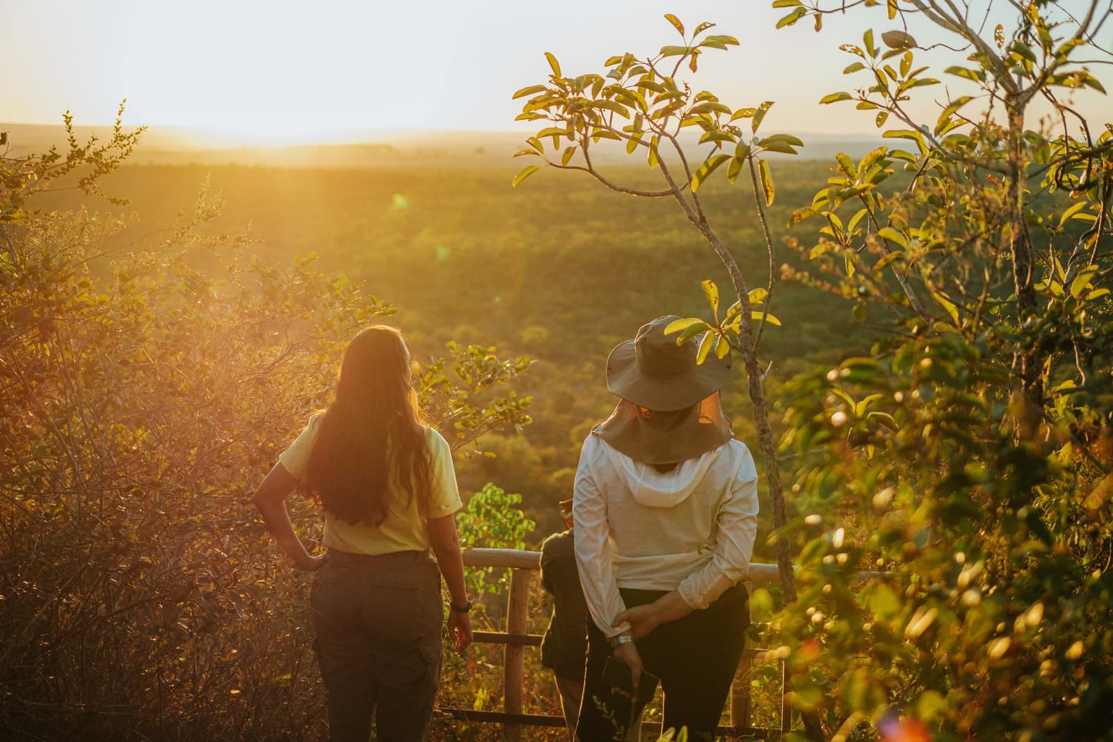 Tem hora que o cerrado pede silêncio e o sol faz o resto. Duas mulheres apoiadas em uma grade de madeira no mirante da Pousada Trijunção, contemplando a vasta paisagem dourada do Cerrado ao pôr do sol
