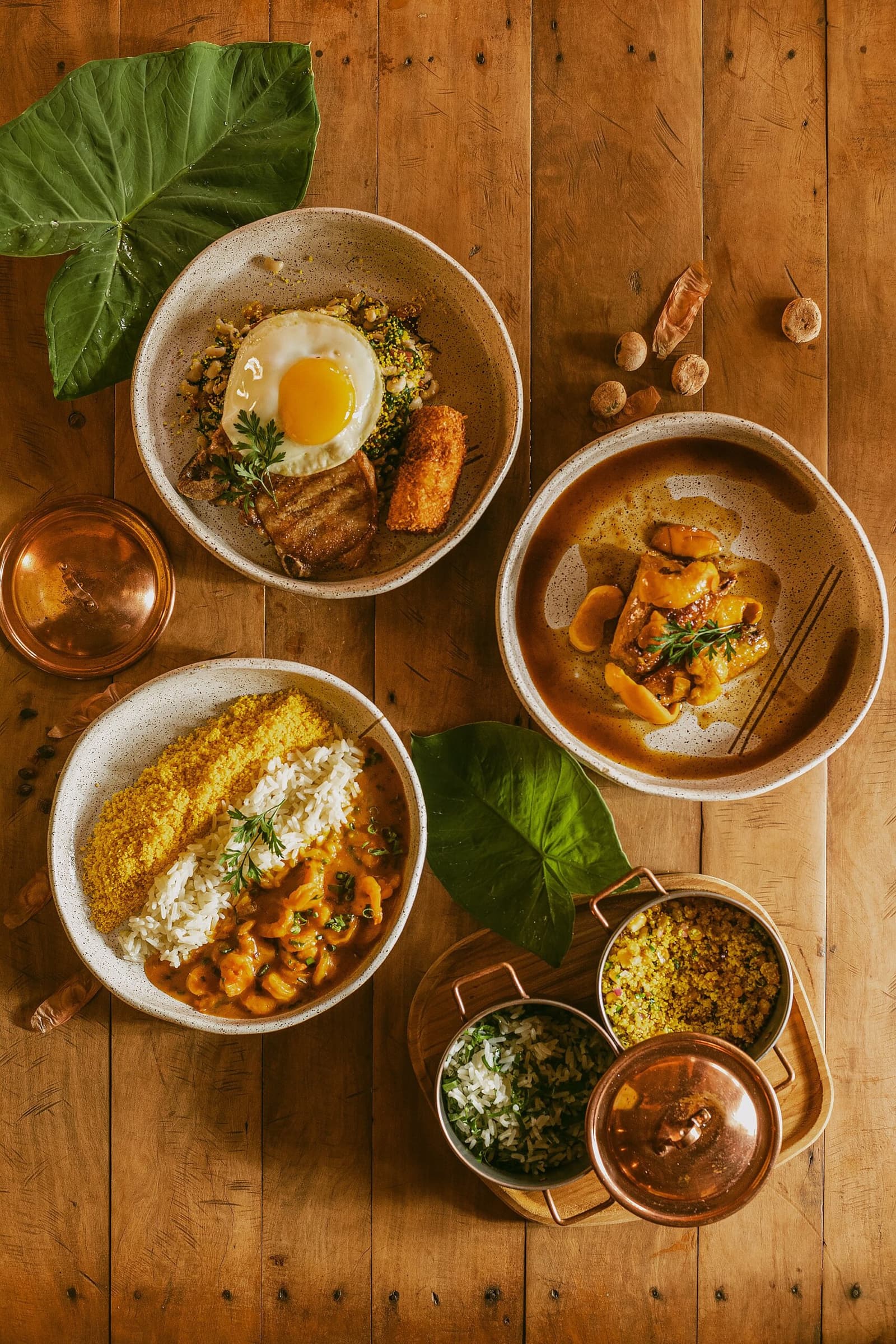 Almoço típico que convence qualquer um a tirar uma soneca depois. Quatro tigelas de cerâmica com pratos típicos brasileiros servidos na Pousada Trijunção: moqueca de camarão com arroz e farofa, bowl de arroz com ovo frito e barriga de porco crocante, ensopado de carne e panelinha de cobre com acompanhamentos sobre uma mesa de madeira