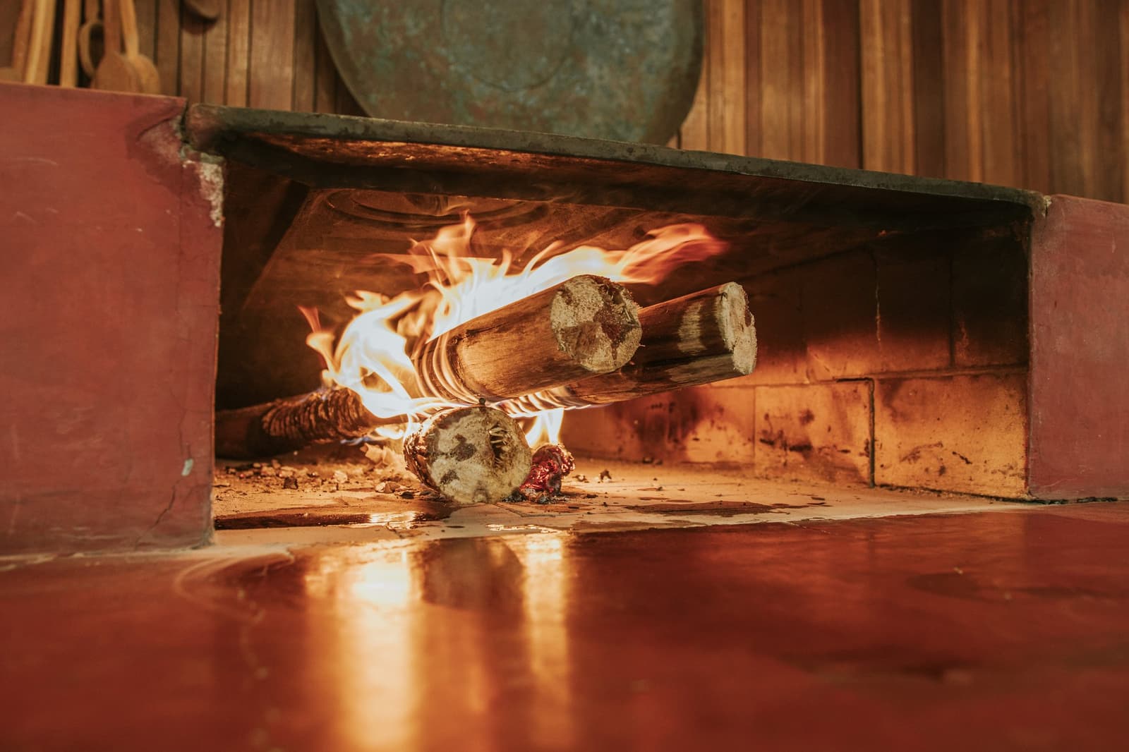 É aqui que começa o sabor, bem antes do prato chegar. Troncos de madeira queimando com chamas vivas em uma fornalha tradicional de pedra na Pousada Trijunção, com o reflexo alaranjado do fogo no piso vermelho polido