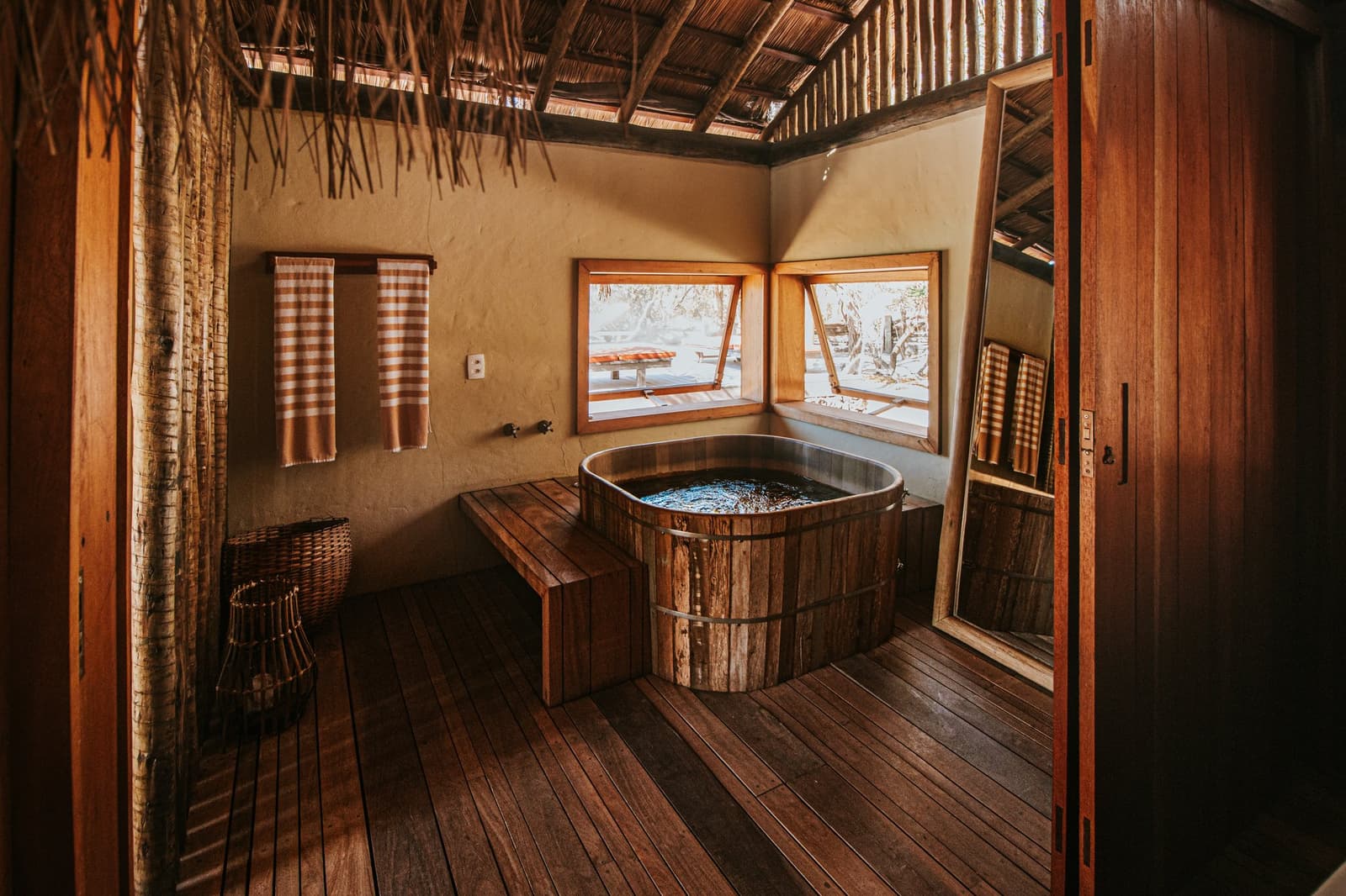 Um banho de banheira no meio do Cerrado. Nem todo conforto é de cidade. Banheira de barril de madeira rústica em uma suíte da Pousada Trijunção, com teto de palha, piso de madeira aquecido e janelas abertas com vista para o Cerrado
