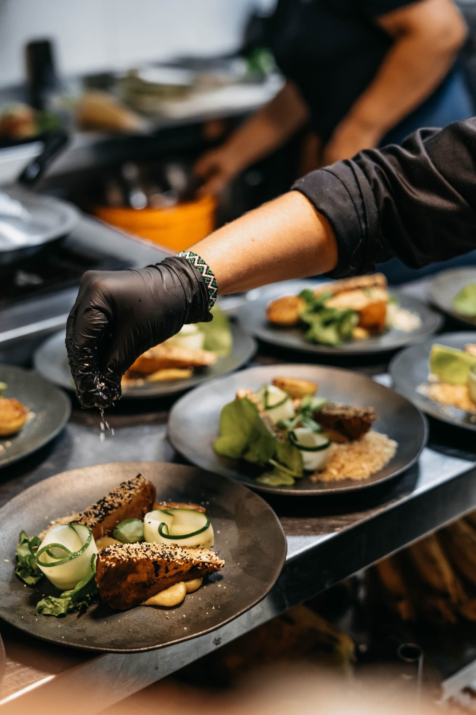 La précision du chef ne faiblit pas, même au fil de l'Amazone. Un chef portant des gants noirs parsème des assaisonnements sur plusieurs assiettes raffinées garnies de portions à croûte de sésame, de concombre en spirale et de verdures fraîches, préparées à bord de l'Untamed Amazon Cruise