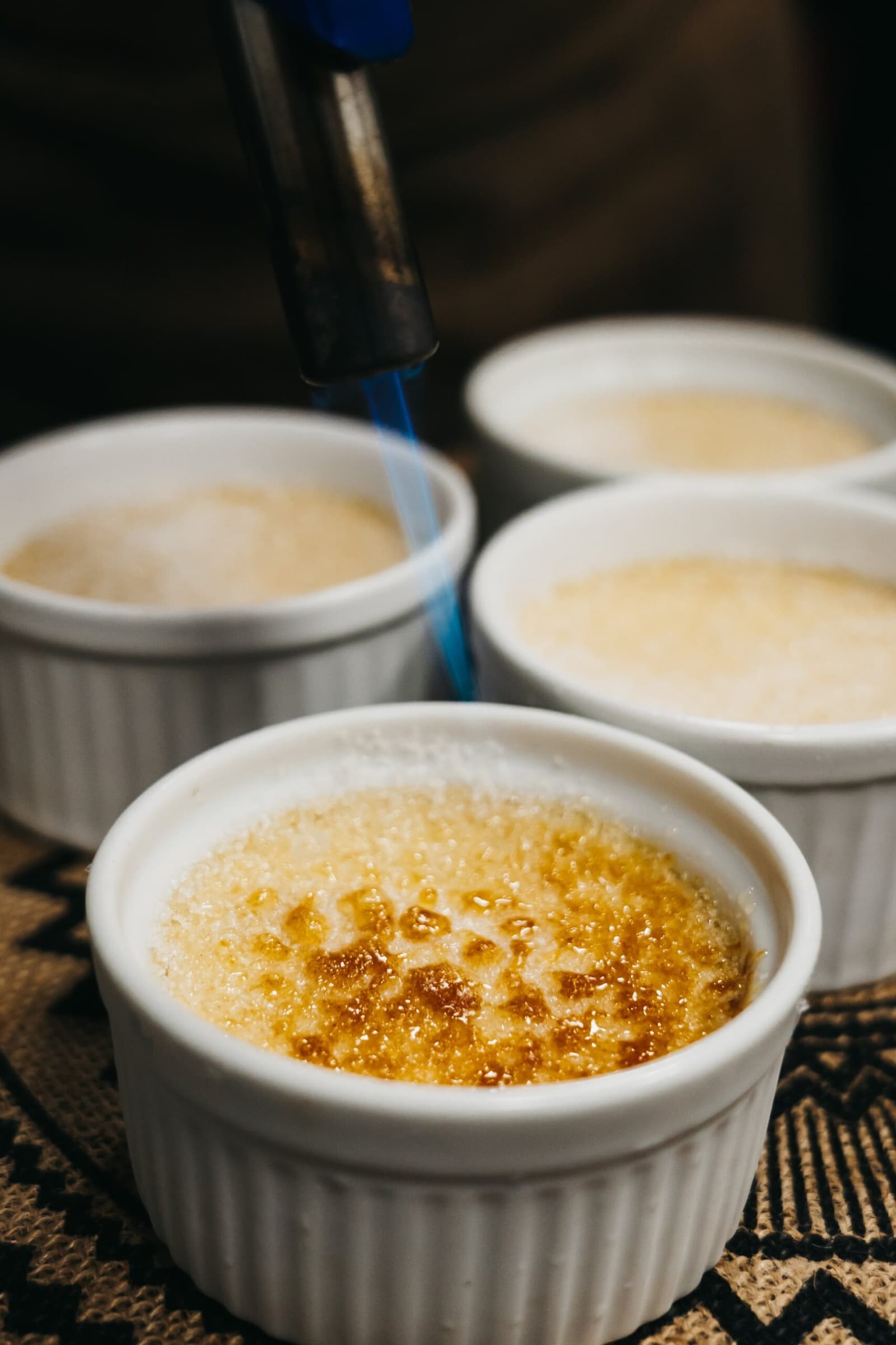 La crème brûlée n'attendait pas d'être en France pour faire son effet. Un chalumeau de cuisine caramélise la croûte de sucre d'une crème brûlée dans un ramequin blanc, avec trois autres ramequins encore non caramélisés en arrière-plan