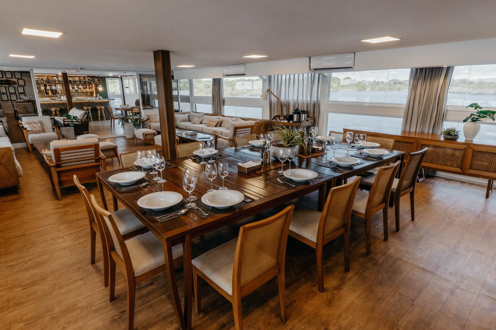Dîner les yeux dans le fleuve, c'est difficile de faire mieux. Table de salle à manger élégamment dressée avec de la porcelaine blanche, des verres à vin en cristal et des chaises en bois à bord de l'Untamed Amazon Cruise, avec vue panoramique sur le fleuve à travers de larges fenêtres et un bar-salon visible en arrière-plan