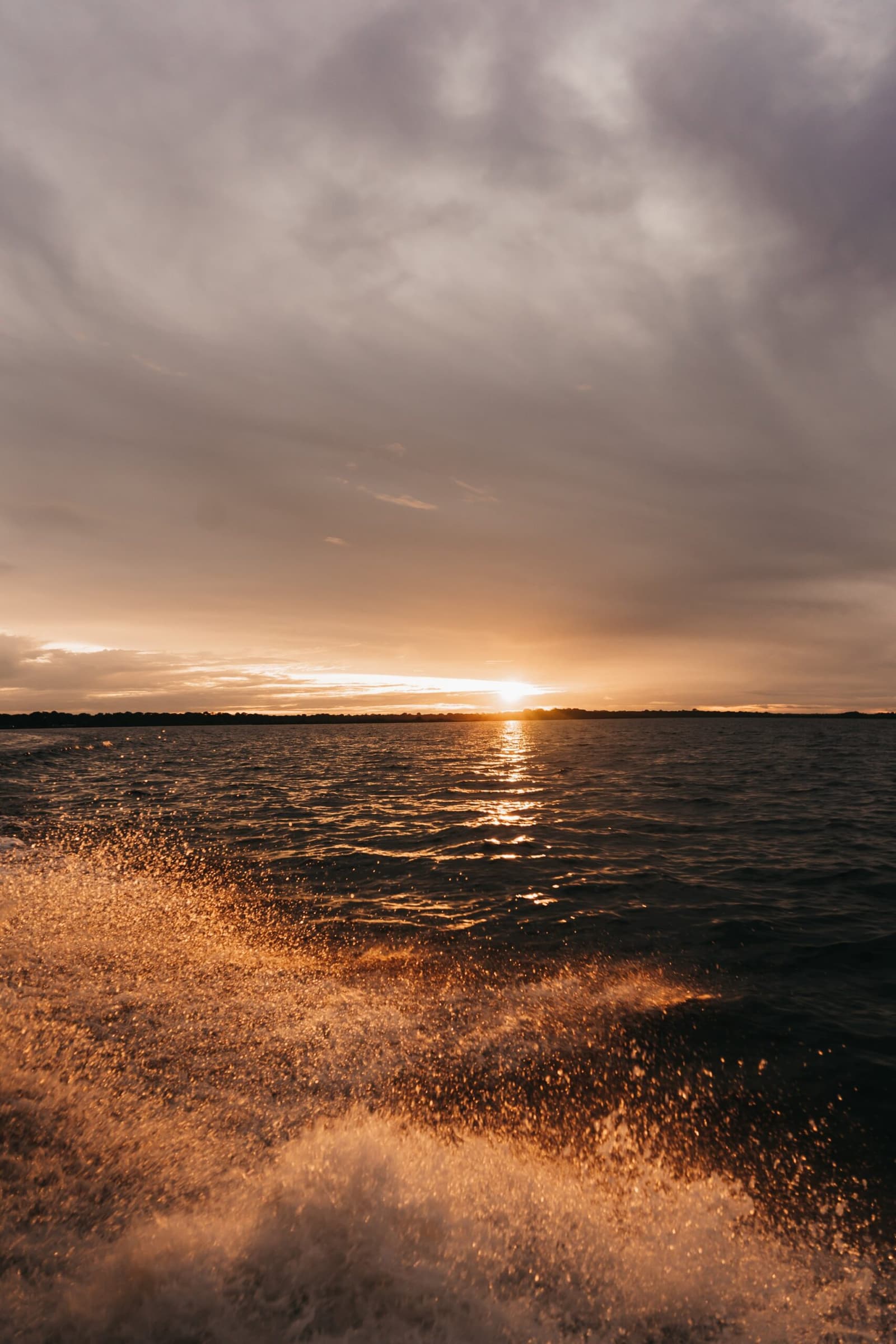 Dernière heure sur l'eau, quand le fleuve prend des airs de métal en fusion. Le sillage doré du bateau scintille au premier plan tandis que le soleil se couche sur un large fleuve lors d'une croisière amazonienne sauvage, avec une ligne d'arbres lointaine sous un ciel nuageux dramatique