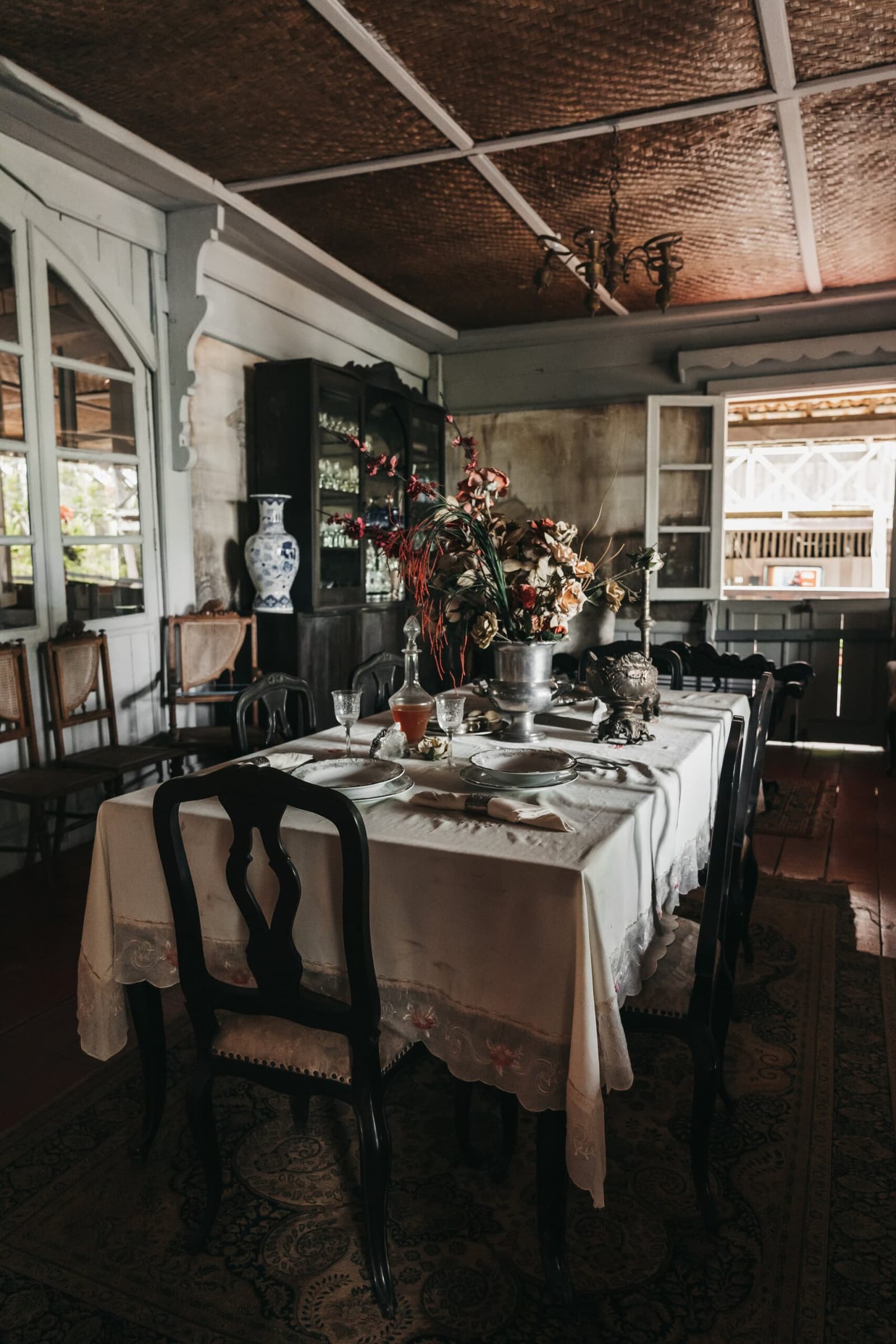 On dirait que les convives du siècle dernier n'ont jamais vraiment quitté la table. Table à manger formellement dressée avec nappe en lin blanc, chaises sculptées sombres, argenterie ancienne, verres en cristal et un centre de table en fleurs séchées dans une salle de style colonial au plafond tressé et à l'armoire à vaisselle d'époque