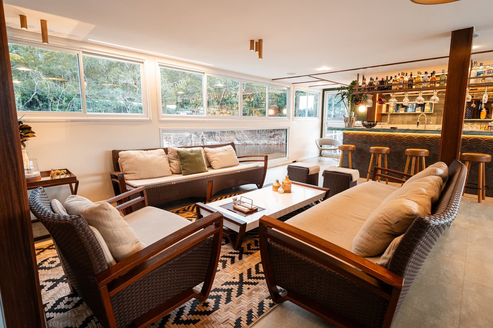 Le bar est là, la forêt aussi. Difficile de choisir où regarder. Le salon à bord de l'Untamed Amazon Cruise avec fauteuils en rotin tressé, coussins crème, tapis à motifs géométriques et un bar entièrement approvisionné, baigné de lumière naturelle sur la forêt fluviale