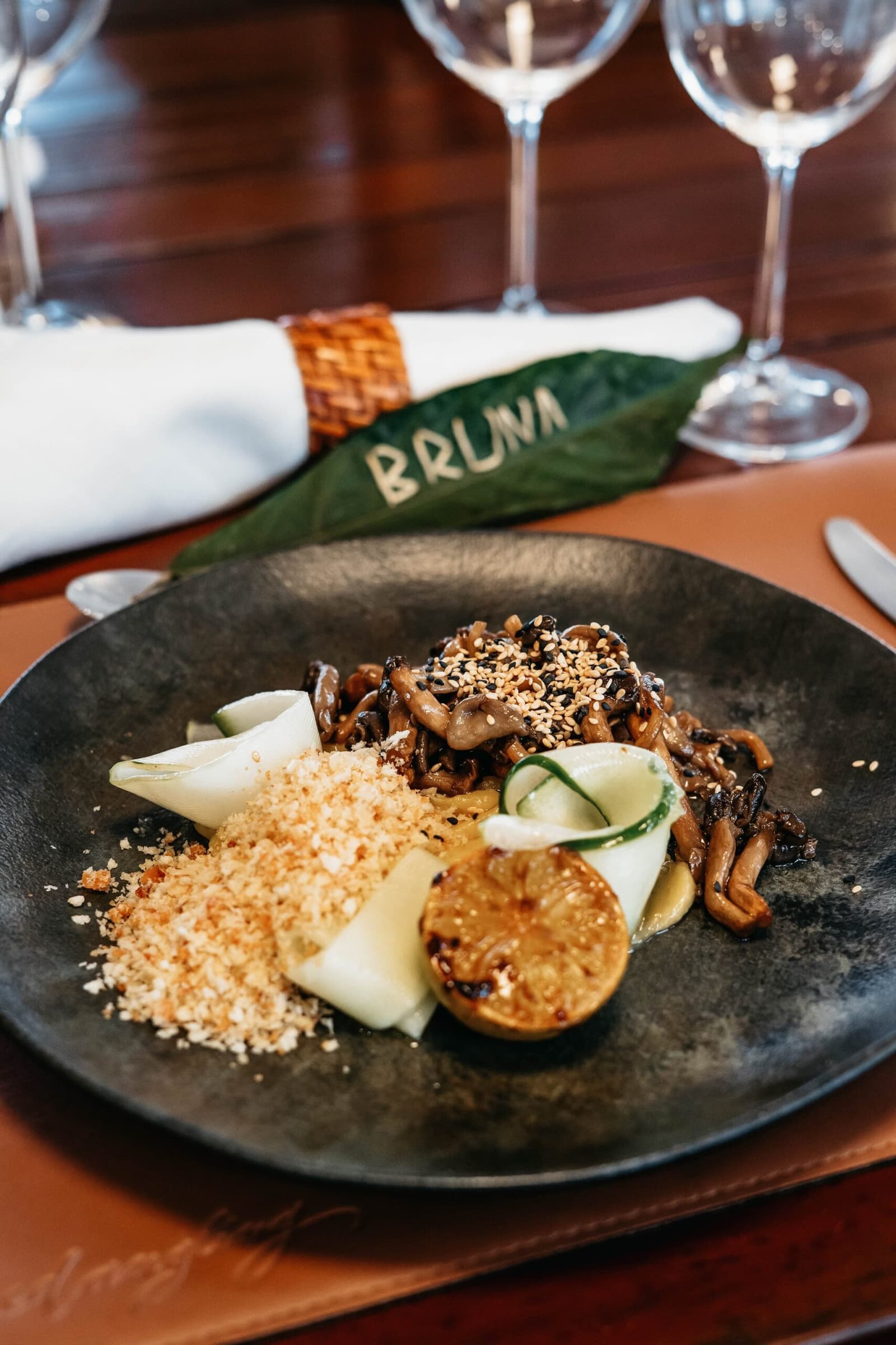 La forêt dans l'assiette, le fleuve à deux mètres. Un plat de champignons sautés mélangés aux graines de sésame, légumes marinés, chapelure dorée et tranche de légume caramélisée, présenté dans une assiette en pierre noire à bord de l'Untamed Amazon Cruise, avec une serviette à anneau tressé et un marque-place en feuille portant le prénom 'Bruna'