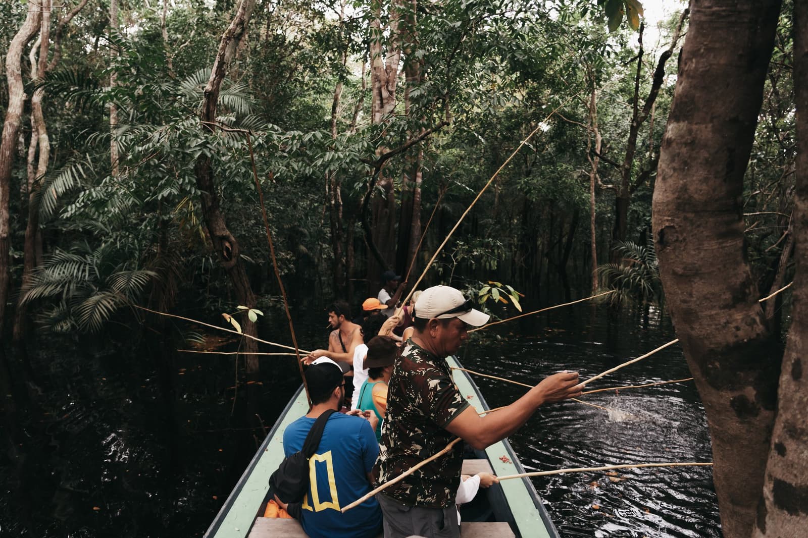La compétition s'installe naturellement dès que les lignes touchent l'eau. Un groupe de voyageurs pêche à la canne en bambou depuis un étroit canot lors d'une croisière Untamed Amazon, entouré d'une forêt igapó inondée et d'eaux noires profondes