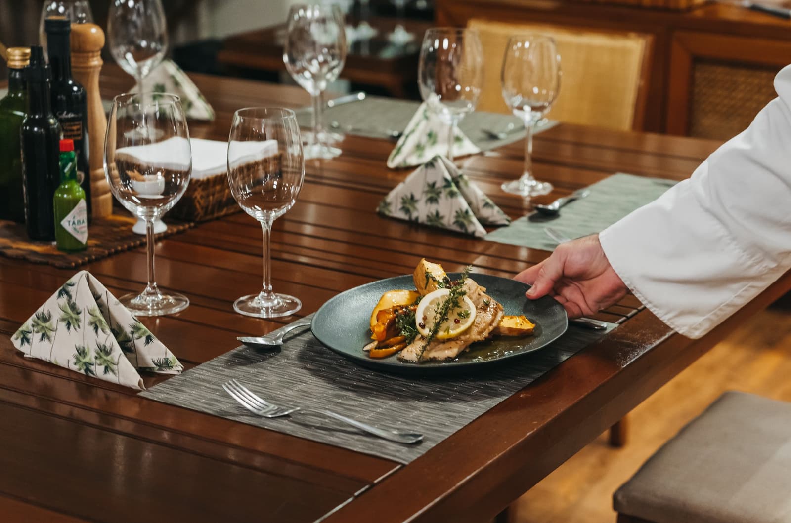 La cuisine soignée fait aussi partie du voyage. Un chef en veste blanche dépose une assiette sombre de poisson grillé avec poivrons rôtis, citron et herbes fraîches sur une table en bois formellement dressée à bord de l'untamed amazon cruise