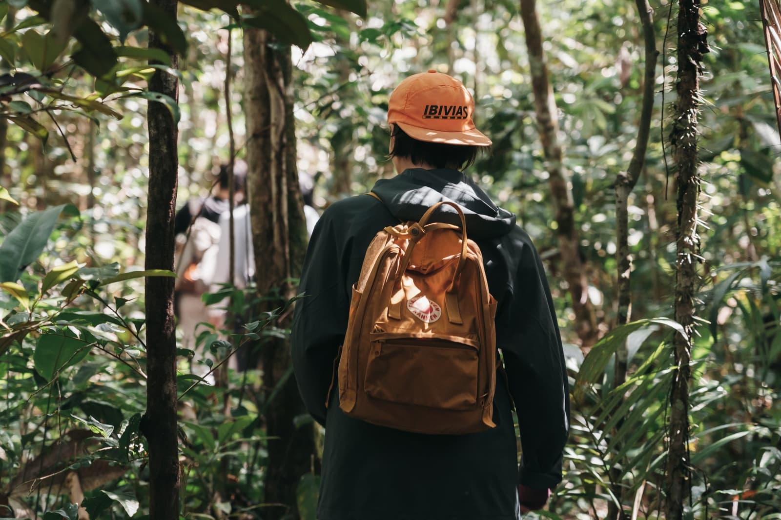 La forêt mène la danse, vous n'avez qu'à suivre. Un randonneur coiffé d'une casquette orange et portant un sac à dos camel traverse une végétation dense lors d'une excursion de l'untamed amazon cruise, avec d'autres voyageurs visibles devant lui sur le sentier