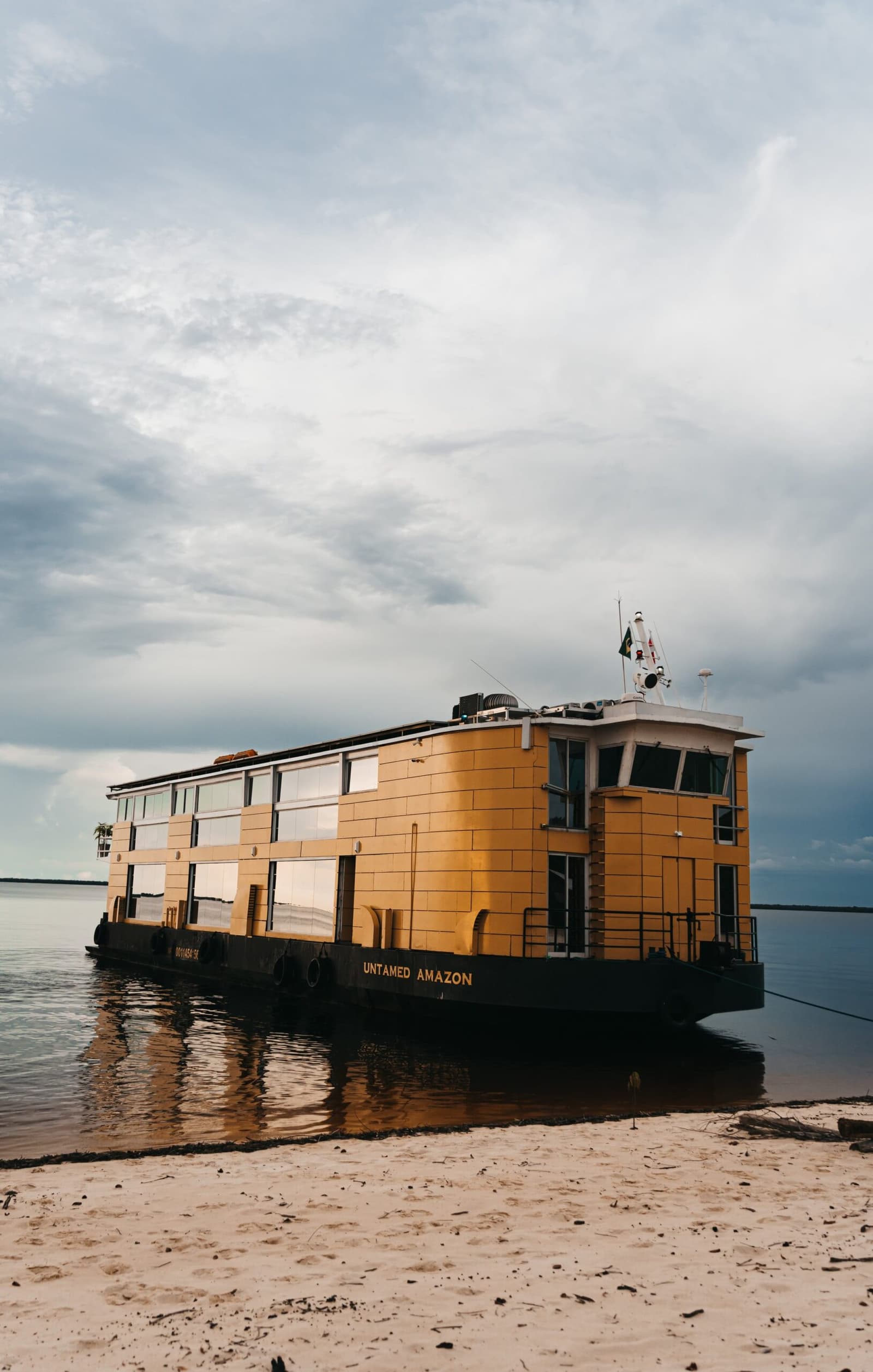 Le camp de base flottant vous attend, amarré au bord du fleuve. Le bateau de croisière Untamed Amazon, peint en jaune doré, amarré sur une plage de sable au bord de l'Amazone sous un ciel nuageux