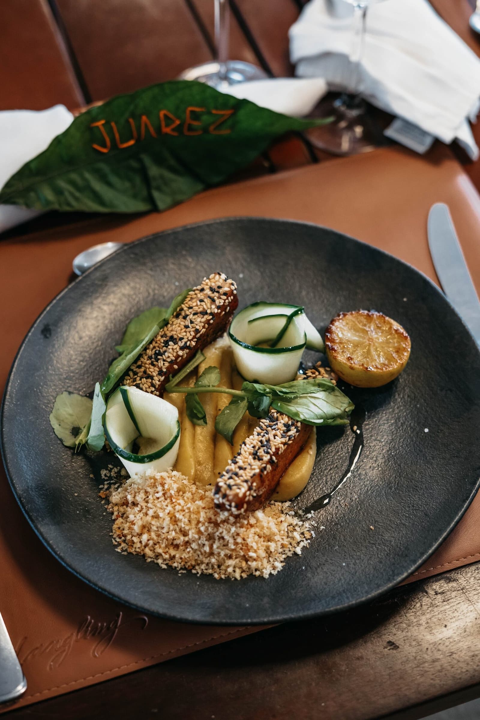 Un dîner qui prouve que la forêt inspire bien au-delà du paysage. Lanières de protéine enrobées de sésame, rouleaux de concombre, purée dorée, chapelure et demi-citron grillé dressés sur une assiette ardoise sombre à bord de l'untamed amazon cruise, avec une feuille marquée 'Juarez' en arrière-plan