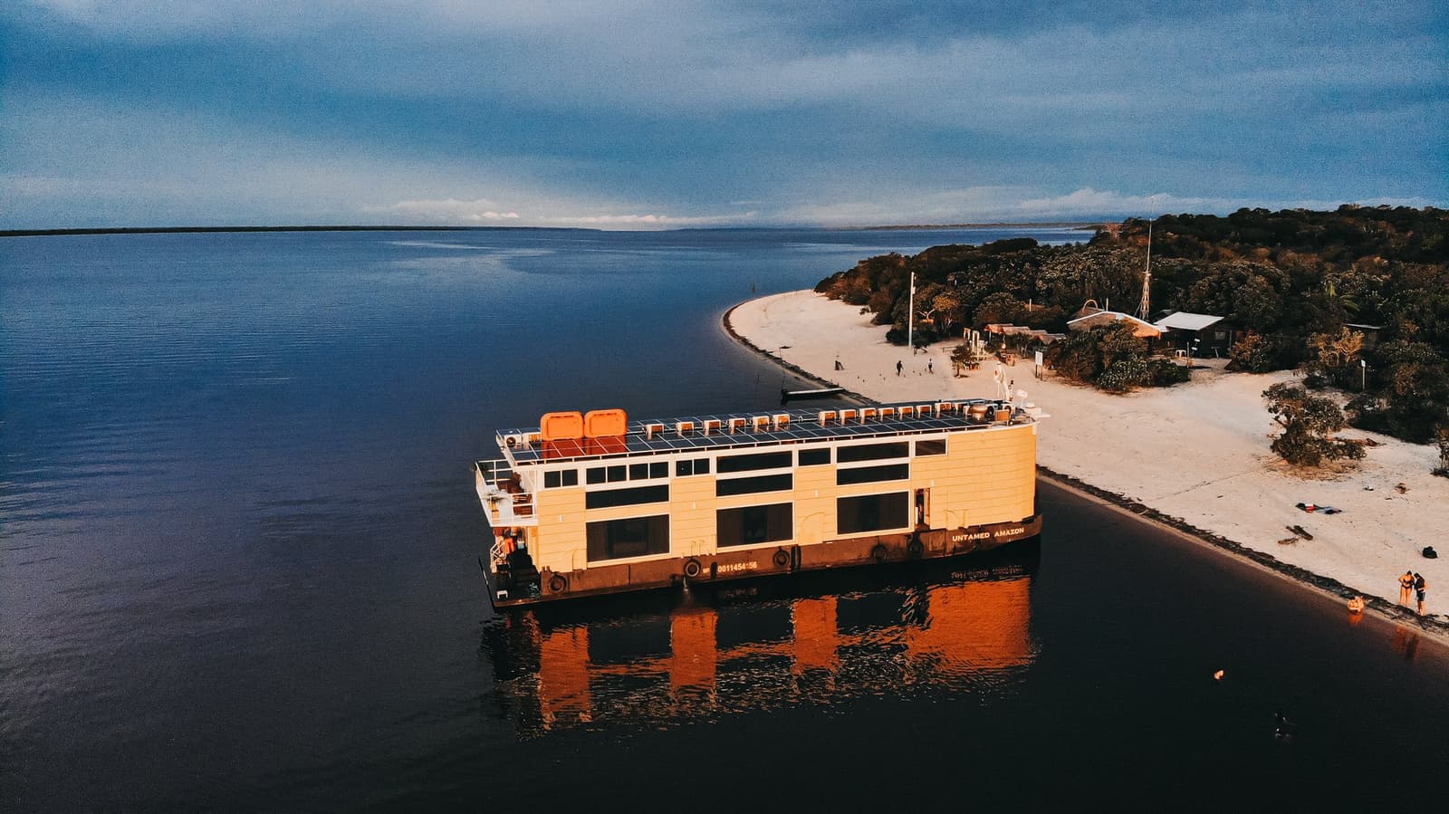 Cabine avec vue sur le fleuve, sans murs ni climatisation forcée. Vue aérienne du bateau solaire de l'Untamed Amazon Cruise amarré contre une plage de sable blanc au crépuscule, avec l'eau sombre et une rive boisée en arrière-plan