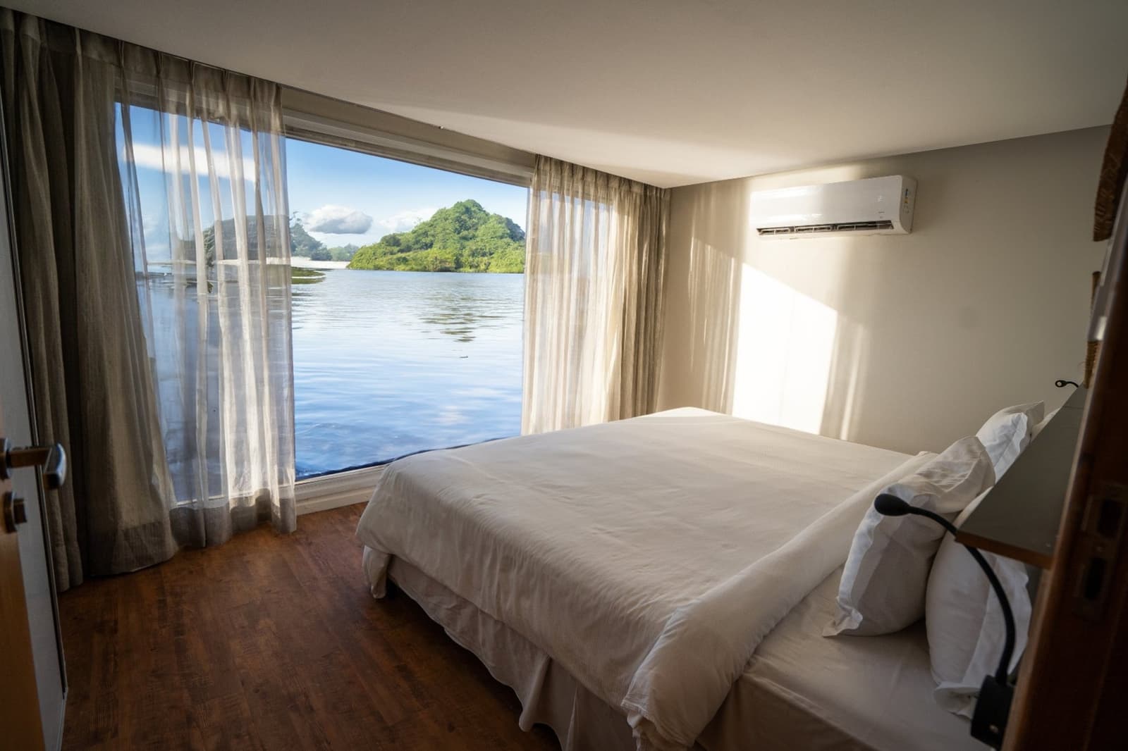 Le fleuve comme fond d'écran, pour de vrai. Cabine climatisée de la croisière Untamed Amazon avec un lit double en linge blanc, parquet en bois et baie vitrée ouvrant sur un fleuve calme et une île boisée