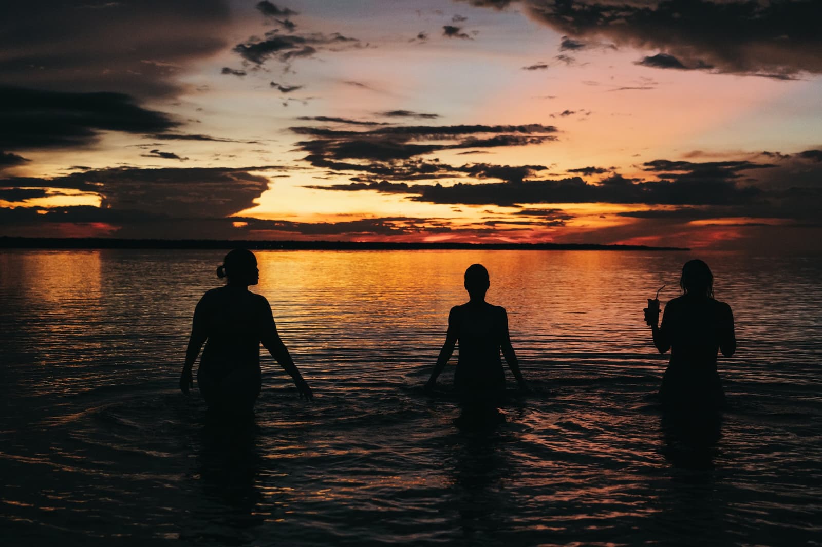 Le fleuve garde la lumière du soir bien après que le ciel l'a lâchée. Trois voyageurs en silhouette jusqu'à la taille dans un fleuve calme sous un coucher de soleil orange et rouge intense lors d'une croisière en Amazonie