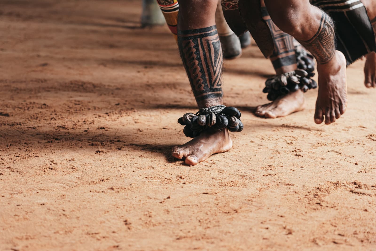Dans ce sol rouge, chaque pas résonne comme un souvenir vivant. Gros plan de pieds nus sur une terre rouge, ornés de bracelets de chevilles en graines et de motifs géométriques peints sur les jambes de danseurs indigènes