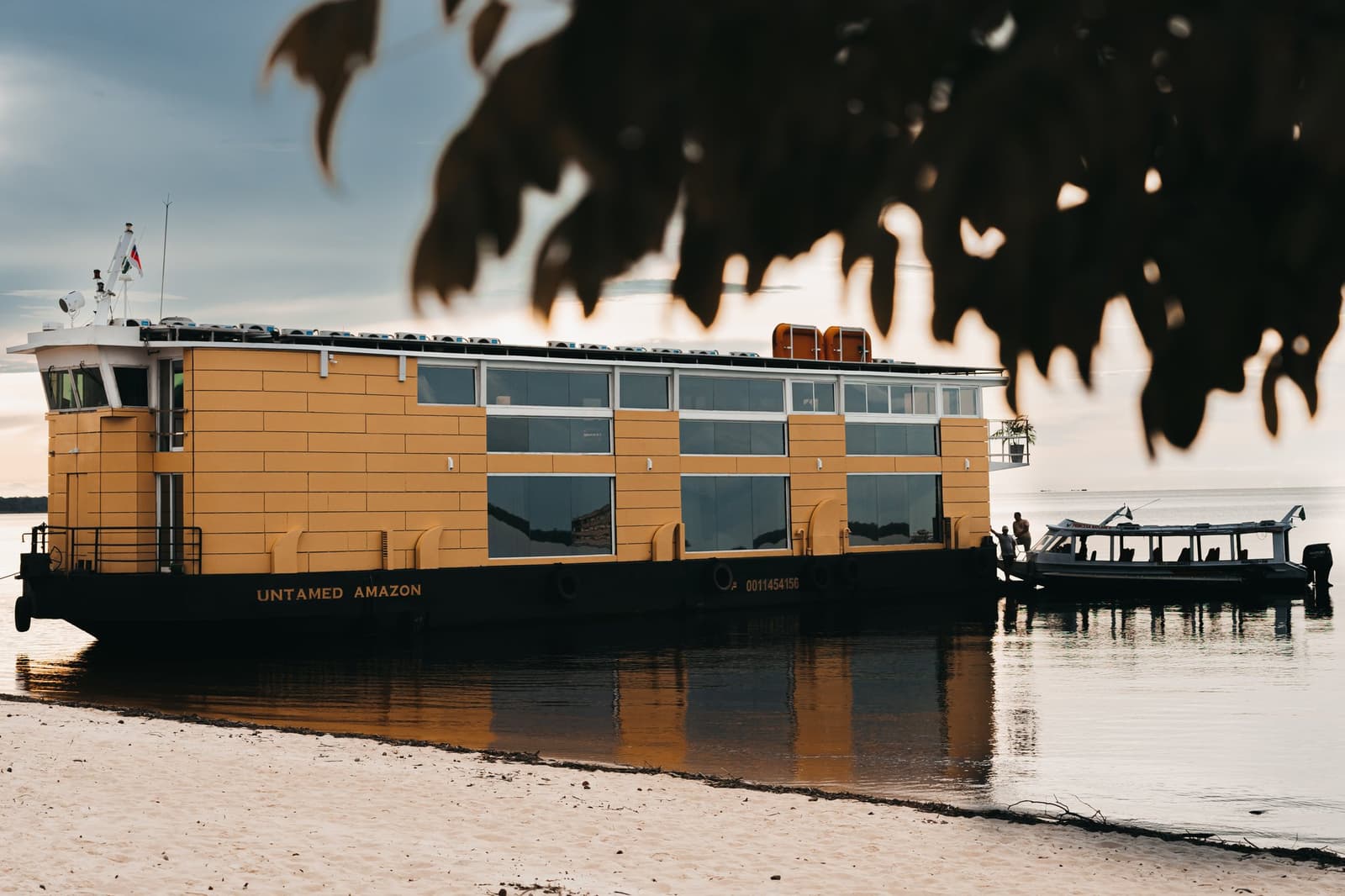 Le camp de base flottant, ancré et prêt à partir. Le navire de croisière Untamed Amazon amarré sur une plage de sable au coucher du soleil, sa coque jaune et ses larges fenêtres de cabine se reflétant dans l'eau calme