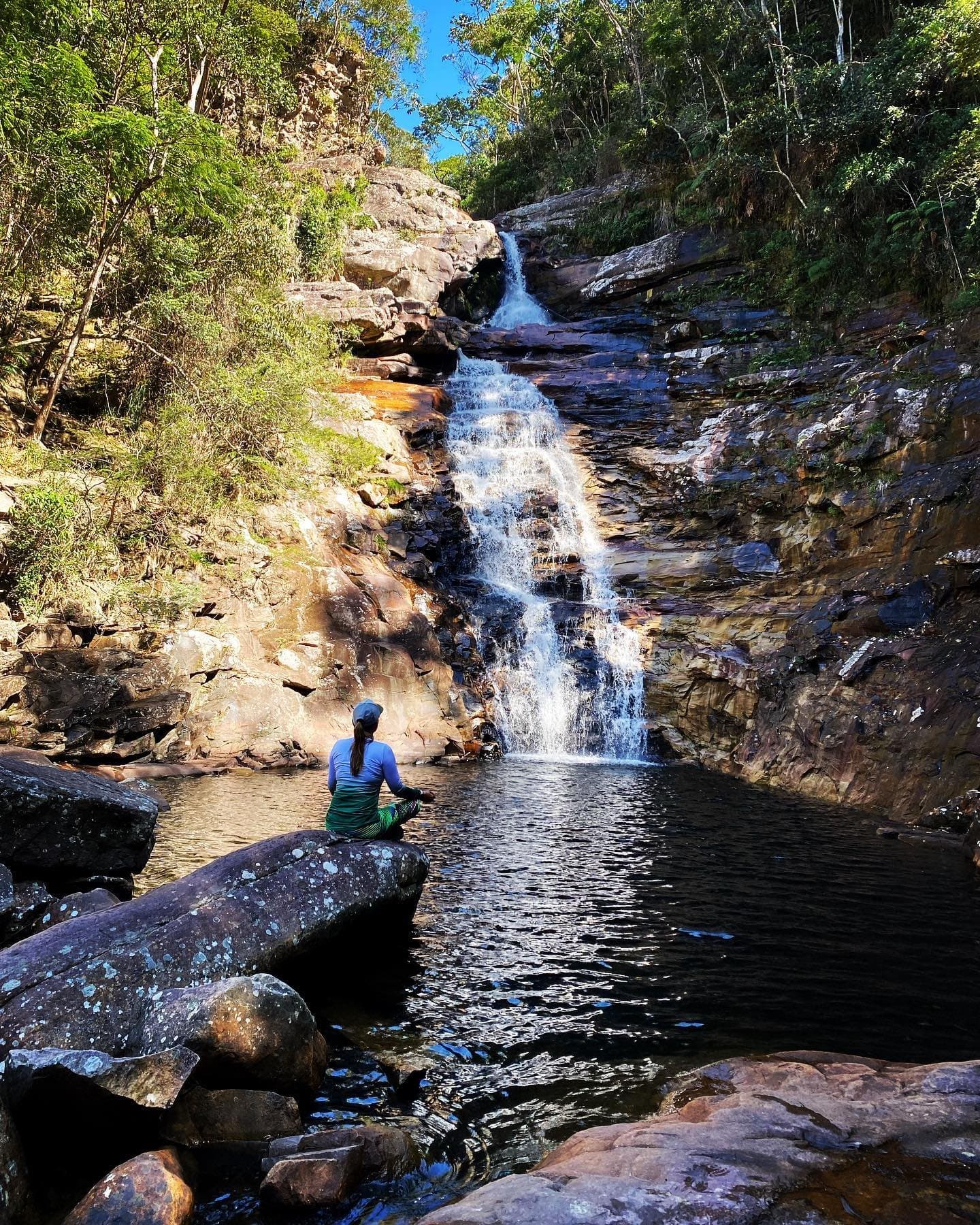 On pose le sac, on s'installe sur un rocher, et on laisse l'eau parler. Une randonneuse assise sur un rocher au bord du bassin naturel d'une cascade en gradins qui dégringole sur des falaises de grès striées de fer lors du trek Vale do Pati, entourée d'une forêt atlantique verdoyante sous un ciel bleu