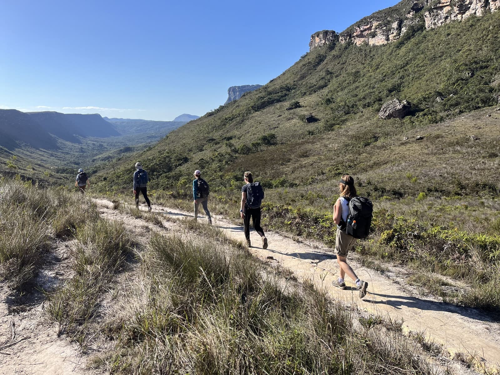 La vallée se dévoile kilomètre après kilomètre, et ça vaut chaque effort. Cinq randonneurs avec sacs à dos marchent en file indienne sur le Vale do Pati Trek, au milieu d'une large vallée bordée de falaises gréseuses sous un ciel bleu limpide