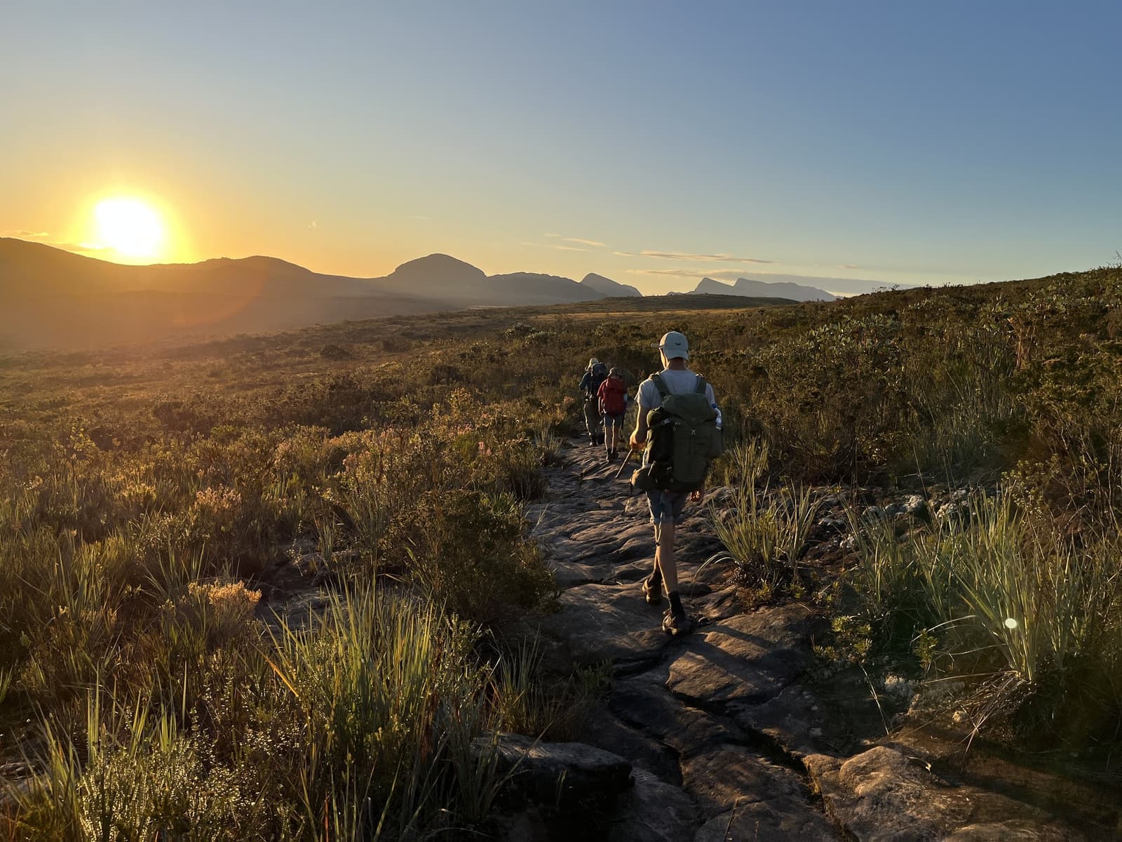 Le soleil à peine levé, les jambes sont déjà en route. Trois randonneurs progressant sur un sentier rocheux à travers la garrigue du Vale do Pati Trek tandis qu'un grand soleil doré se couche derrière une crête montagneuse lointaine