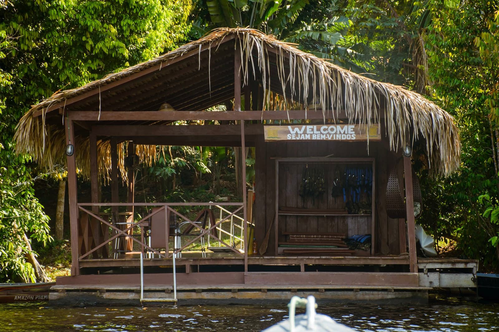 Per boot aankomen bij een bordje in twee talen, dat is een goede start. Een houten steigerpaviljoen met rieten dak en een 'Welcome / Sejam Bem-Vindos'-bord, gefotografeerd vanaf het water tijdens een Amazon en Pantanal tour, omgeven door dicht regenwoud