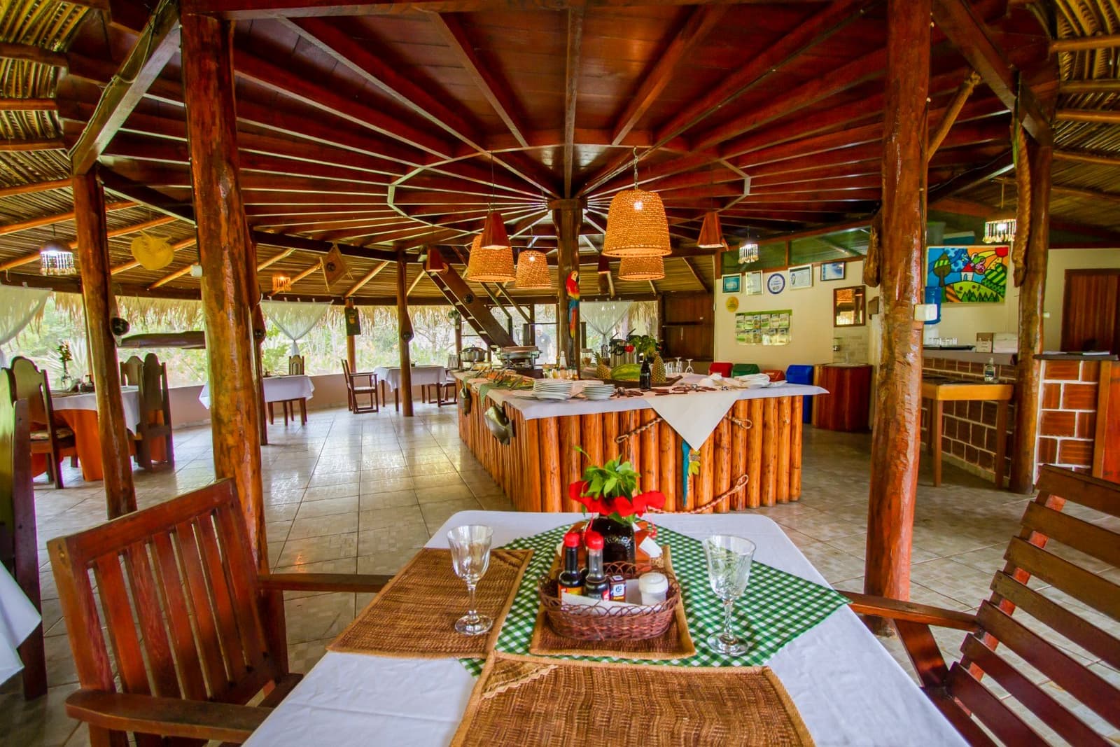 Tafelen met het wetland op een steenworp afstand, zo simpel kan het zijn. Openlucht eetruimte met rieten dak van een Pantanal-lodge op een Amazon- en Pantanal-rondreis, met een rustiek buffet van boomstammen, gevlochten hanglampen, wit gedekte tafels met wijnglazen en een rieten mandje voor condimenten, met uitzicht op het groen