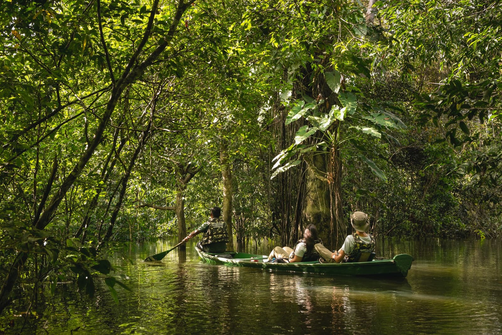 Half ondergelopen bos, zacht geplons, en geen haast om ergens te zijn. Photo: Guilherme Gomes Een gids peddelt twee gasten door een overstroomd woud in een groene kano tijdens een amazon rainforest ecolodge trip in Brazilië, omringd door hoge bomen en breedbladige planten die weerspiegelen in het spiegelstille water