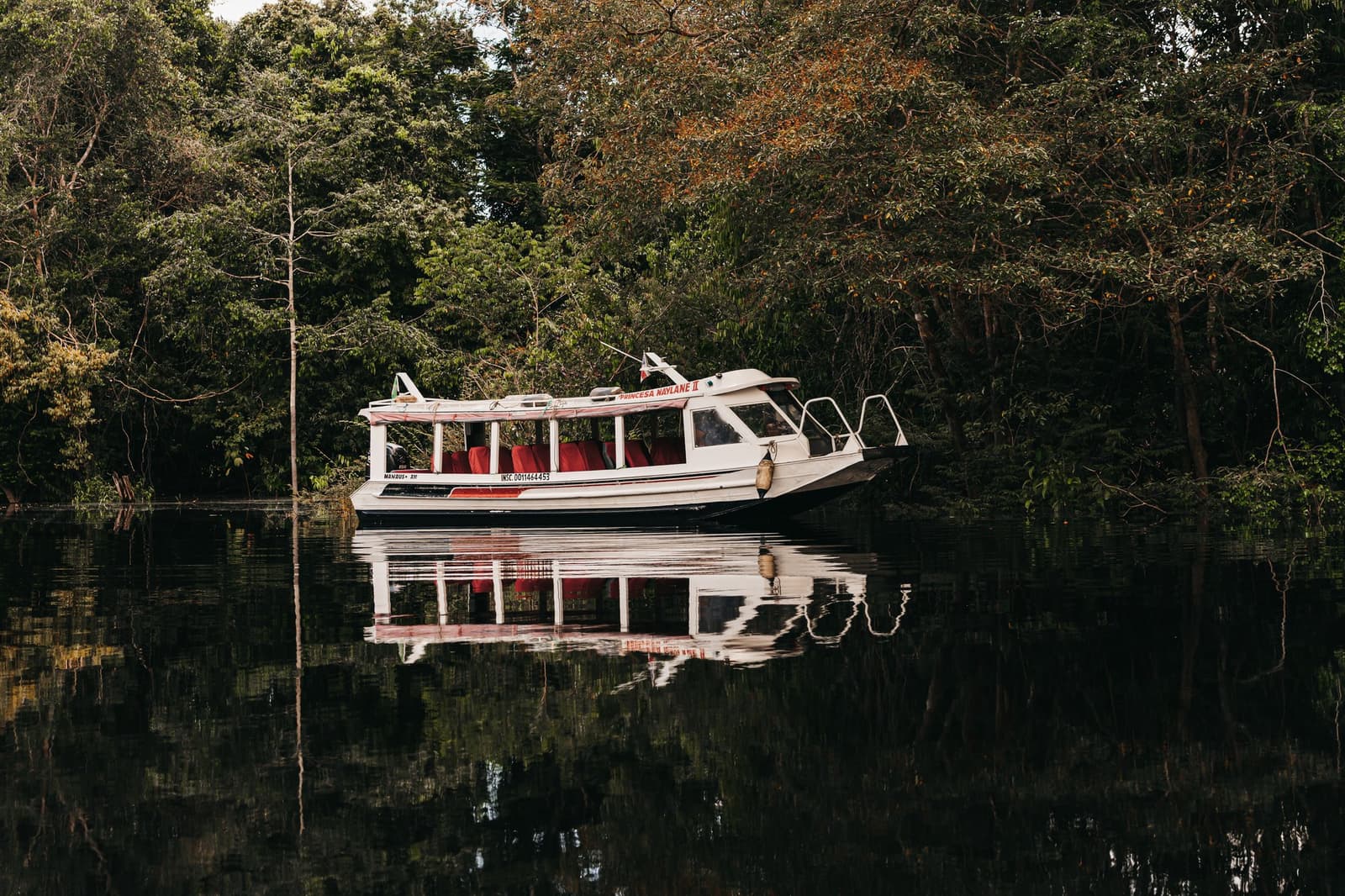De boot ligt klaar, het oerwoud trekt al aan je. Het witte passagiersboot Princesa Naylane II met rode stoelen ligt aangemeerd op donker zwart water, weerspiegeld in het spiegelgladde oppervlak, omringd door het dichte bladerdak van het Amazonewoud.