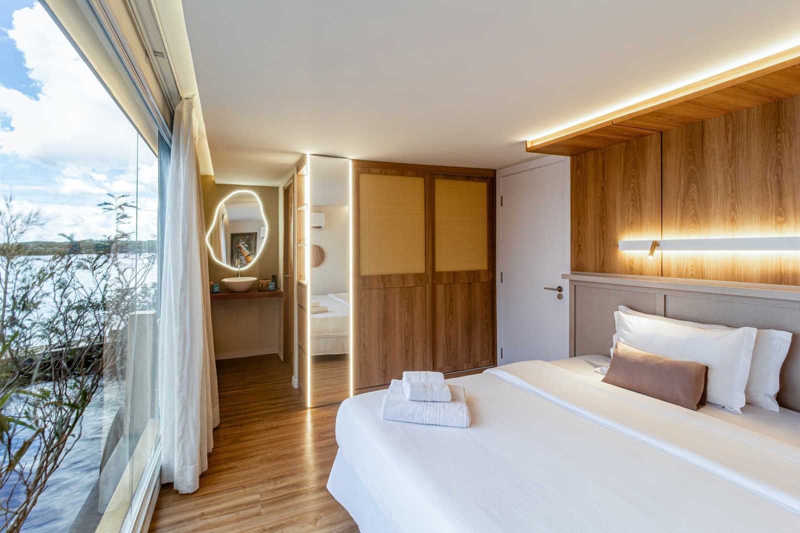 Comfortabel slapen terwijl de rivier langs glijdt. Photo: STUDIO CREATE Lichte suite op een luxe Amazone-cruise in Brazilië, met een tweepersoonsbed met witte beddengoed, een warme houten hoofdeinde met LED-verlichting, vloer-tot-plafondvensters met uitzicht op een brede rivier en een open ruimte met een verlichte kaptafelsspiegel