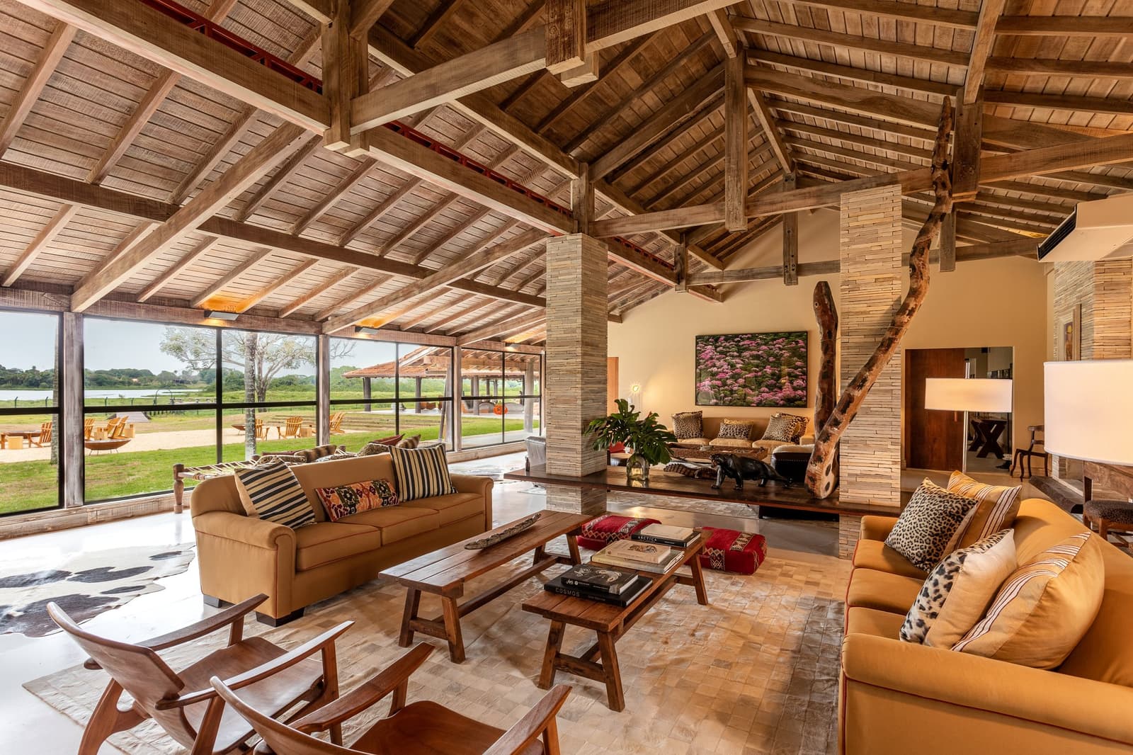 Uma sala que convida a ficar, mesmo sabendo que os onças andam lá fora. Lounge aberto do lodge de Caiman em uma viagem de luxo ao Pantanal e Amazônia, com vigas de madeira aparente no teto, sofás cor de âmbar com almofadas de estampa animal, uma escultura de madeira flutuante e janelas do chão ao teto com vista para as planícies alagadas