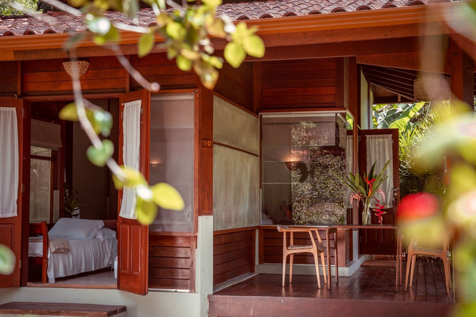 Dschungel draußen, weiches Leinen drinnen, so soll es sein. Photo: Samuel Melim Holzbungalow Nummer 10 auf einer Luxury-Pantanal-und-Amazonas-Reise, mit offenen Fensterläden, die ein weißes Bett enthüllen, einer schattigen Veranda mit Holzstühlen und tropischem Grün im Vordergrund