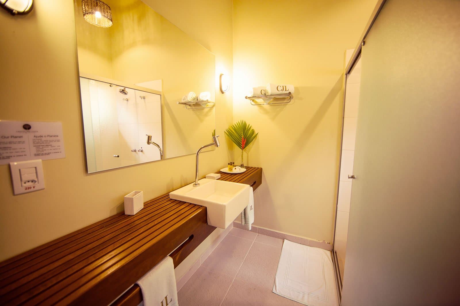 Teakholz, Monogramm-Handtücher, eine Helikonie. Der Dschungel wartet gerne noch ein paar Minuten. Photo: | SamuelMelim Photography Badezimmer einer Lodge auf einer Luxus-Pantanal-und-Amazonas-Reise mit eckigem Aufsatzbecken auf einem Teakholz-Waschtisch, warmgelben Wänden, Handtüchern mit Monogramm und einem tropischen Helikoniastrauß