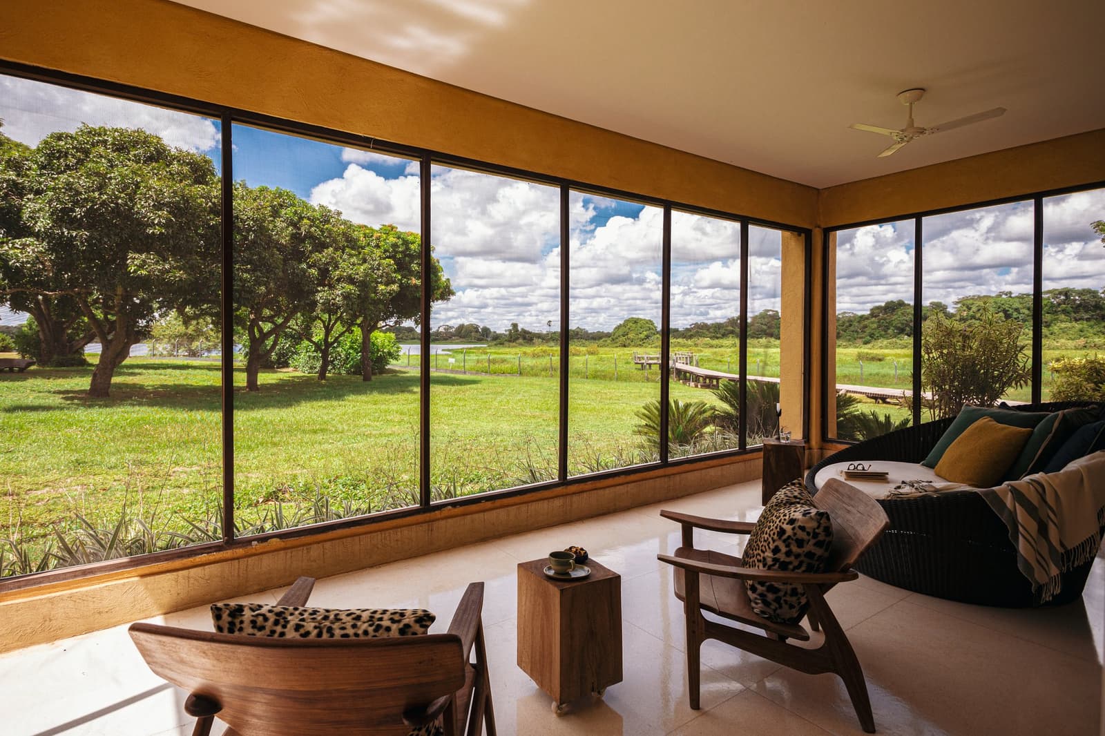 Der beste Platz im Pantanal, mit Deckenventilator und frischem Kaffee. Photo: Felipe Castellari Überdachter Loungebereich einer Luxus-Lodge für eine Pantanal-und-Amazonas-Reise, mit Holzsesseln und tiergemusterten Kissen vor raumhohen Fenstern mit Blick auf eine grüne Überschwemmungsebene, einen Holzsteg und einen bewölkten Himmel