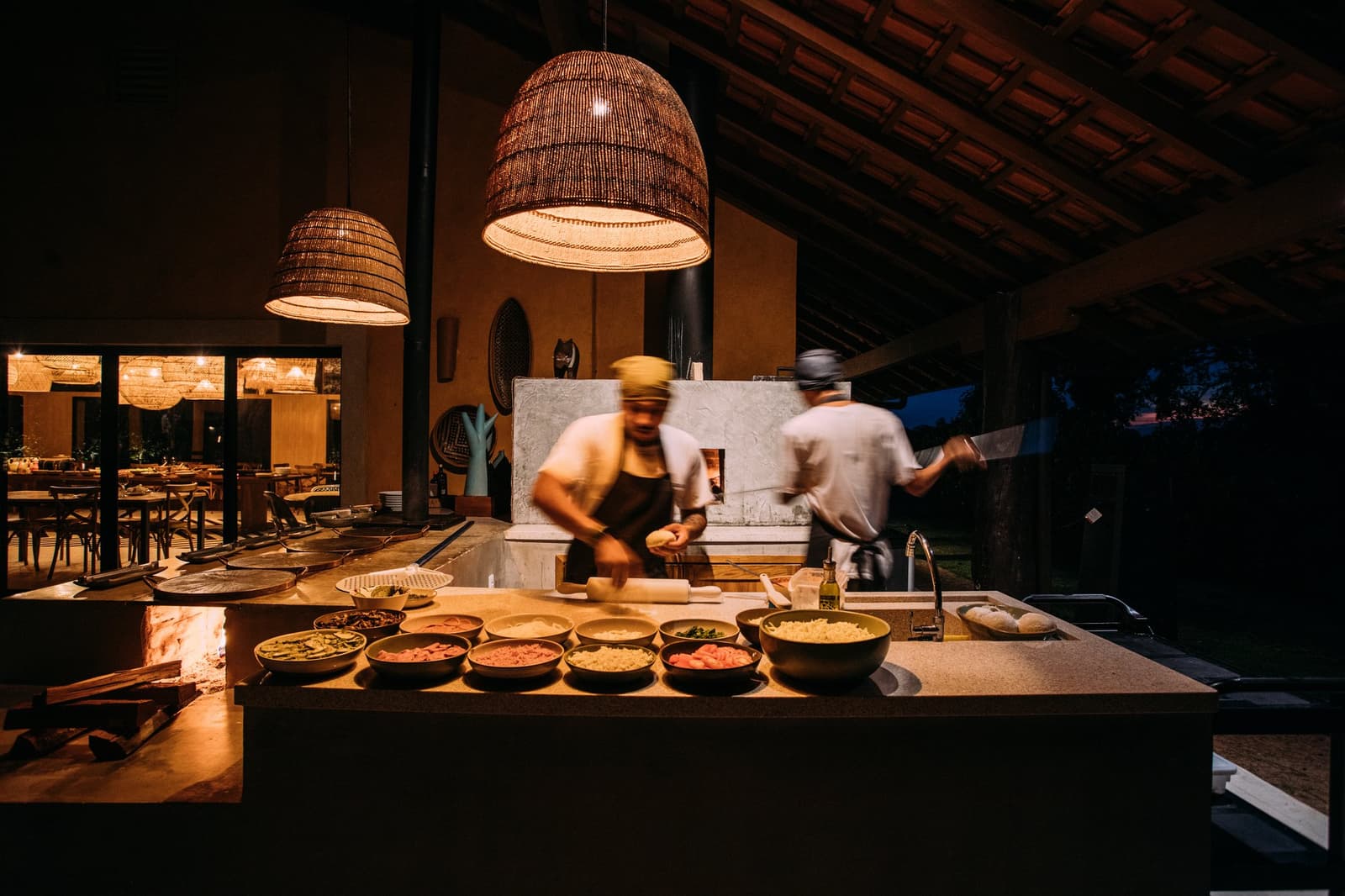 O jantar não aparece do nada, ele nasce bem ali, na sua frente, no ritmo certo. Dois cozinheiros em movimento em uma estação de cozinha aberta com forno a lenha durante uma viagem de luxo pelo Pantanal e Amazônia, com tigelas de ingredientes frescos alinhadas em um balcão de pedra sob luminárias de vime aquecidas ao entardecer