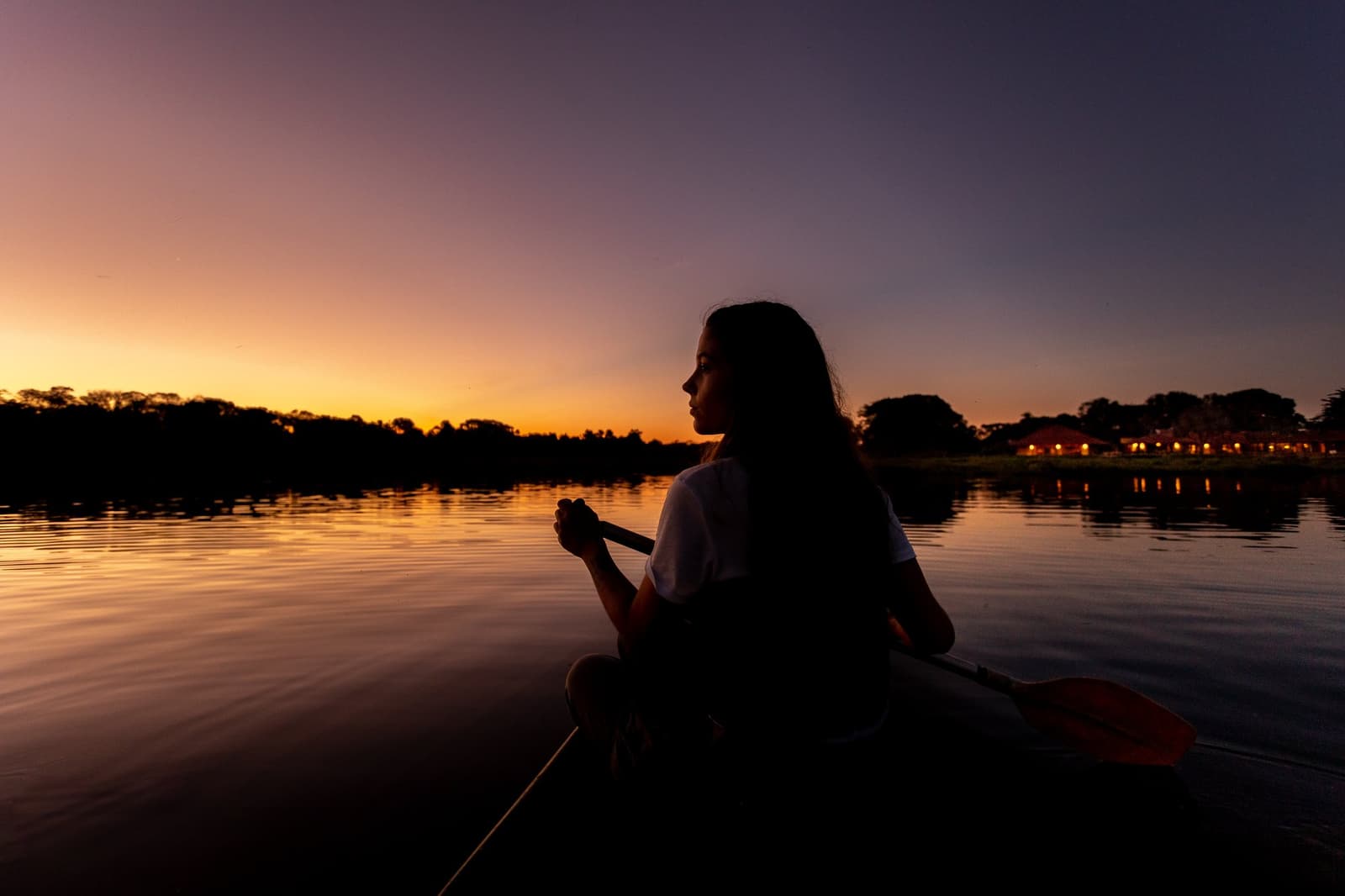 O céu muda de cor em minutos. Bom estar no meio do rio quando isso acontece. Silhueta de uma mulher segurando um remo em um caiaque num rio calmo ao pôr do sol durante uma viagem de luxo ao Pantanal e à Amazônia, com céu laranja e lilás refletido na água parada e as luzes da lodge visíveis na margem oposta