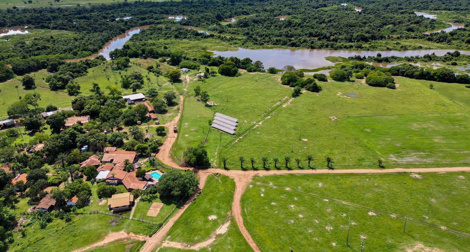 Zonnepanelen naast de veepaden. Eco-lodges in het Pantanal zijn een heel eind gekomen. Luchtfoto van een Pantanal-lodge met zonnepanelen, gebouwen met rode pannendaken, een zwembad en zandpaden door open groene velden naast een kronkelende rivier en galeriewoud
