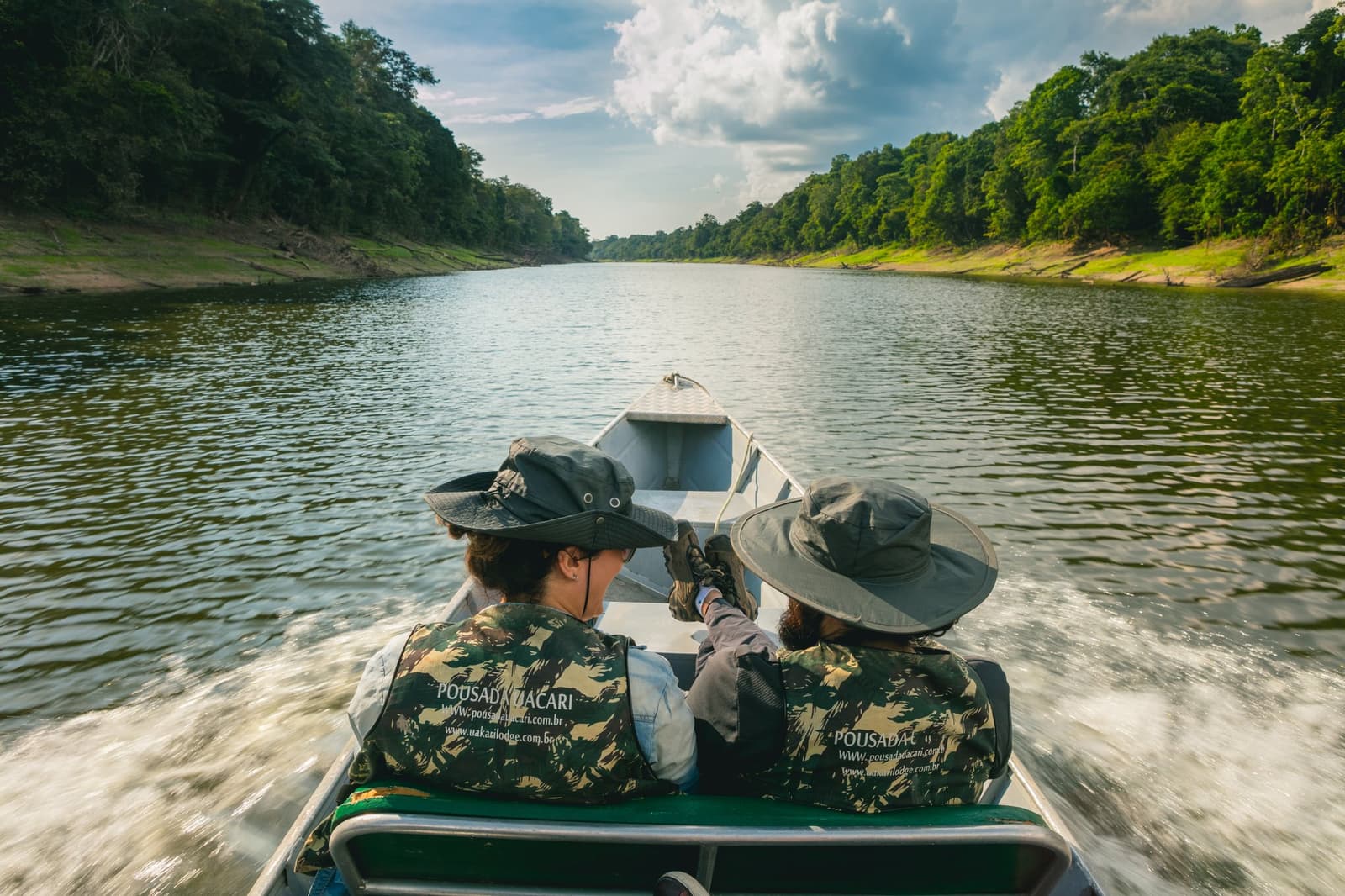 De rivier toont je nooit alles tegelijk. Dat is precies de bedoeling. Photo: Guilherme Gomes Twee gasten in camouflage zwemvesten en brede hoeden zitten aan de boeg van een speedboot op een amazon rainforest ecolodge trip in Brazilië, wijzend naar voren terwijl de boot door een rustige donkere rivier snijdt, omzoomd door dicht oerwoud