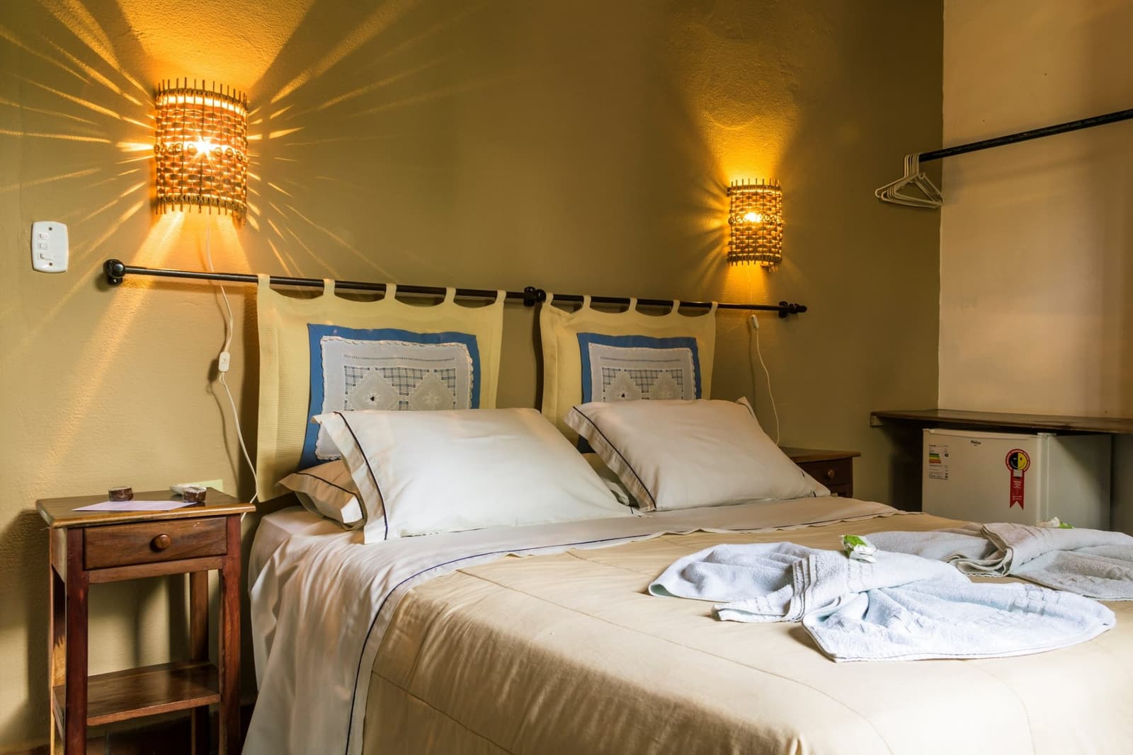 Klein, warm en precies genoeg na een dag vol wilde natuur. Knus tweepersoonskamer in een Pantanal-lodge op de Pantanal Bonito-tour, met warme gevlochten bamboe wandlampen, roomwit beddengoed met blauwe kussensloopaccenten, gevouwen badjassen en een houten nachtkastje