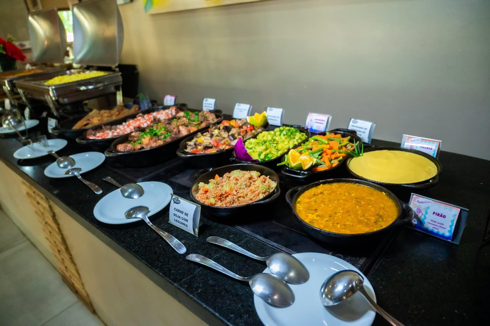Pirão, polenta, carne de soja, de bordjes leggen het uit, de honger doet de rest. Een Braziliaans buffet tijdens een Amazon- en Pantanal-tour, met gelabelde gerechten waaronder pirão-vissoep, polenta, carne de soja com legumes, roergebakken groenten met sinaasappelpartjes en een graansalade, geserveerd in zwarte kleipotten op een donker granieten aanrecht