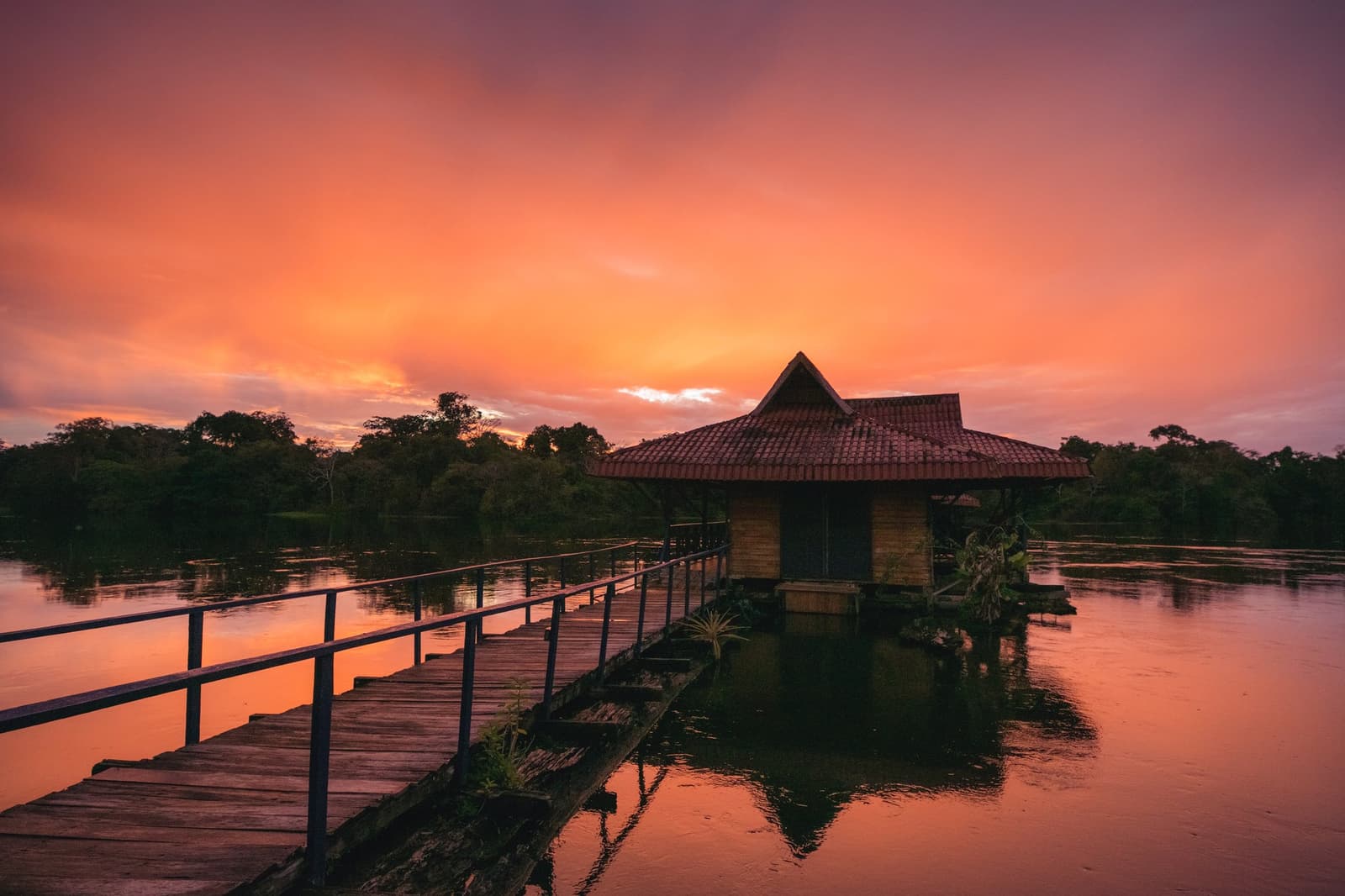 De gedachte die elke avond voor het eten blijft hangen. Photo: Guilherme Gomes Een houten loopbrug leidt naar een bungalow boven het water met een terracotta dak op een rustig bosmeer tijdens een amazon rainforest ecolodge trip in brazilië, de hemel gloeit oranje en roze bij zonsondergang terwijl de boomsilhouetten weerspiegelen in het spiegelgladde water