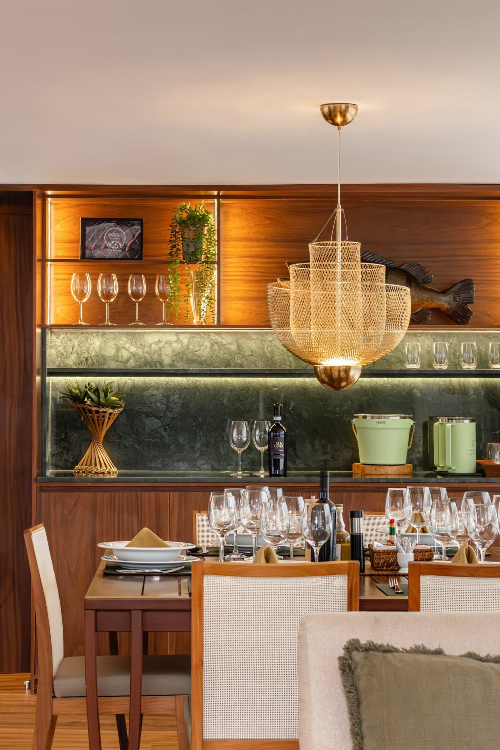 Dineren op de rivier smaakt beter als de tafel er zo bij staat. Photo: STUDIO CREATE Elegant gedekte eettafel aan boord van een luxe Amazone-cruise in Brazilië, met wijnglazen, wit porselein en rieten stoelen voor een verlicht walnotenhouten wandmeubel met glaswerk, een wijnfles en een gesneden houten vissculptuur onder een gevlochten hanglamp