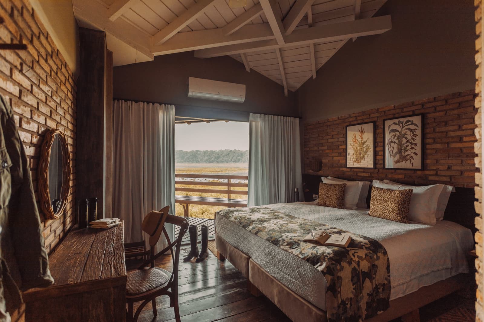 Gummistiefel bereit, Buch aufgeschlagen. Safarialltag, wie er sein sollte. Photo: Felipe Castellari Rustikales Luxus-Doppelzimmer auf einer Luxury Pantanal and Amazon Trip-Reise mit Backsteinwänden, dunklem Holzboden, Leoparden-Kissen, Gummistiefeln an der Balkontür und einem breiten Fenster mit Blick über das Feuchtgebiet
