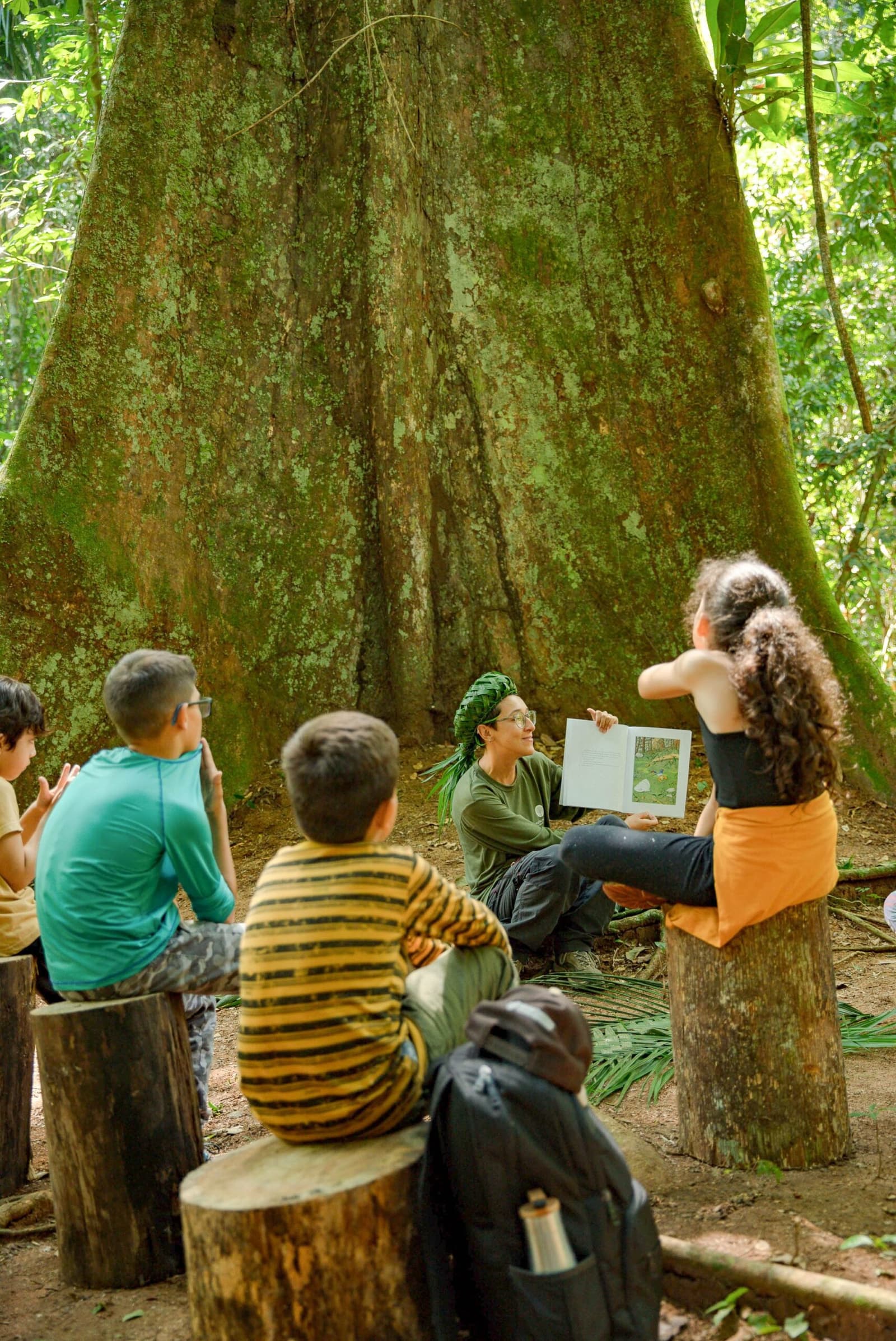 Kein Klassenzimmer, nur ein uralter Baum und echte Neugier. Eine Guides sitzt lächelnd vor den moosbedeckten Wurzeln eines riesigen Regenwaldbiums und liest einer kleinen Kindergruppe auf Baumstümpfen aus einem bebilderten Buch vor — eine Naturaktivität auf einer Luxus-Pantanal-und-Amazonas-Reise