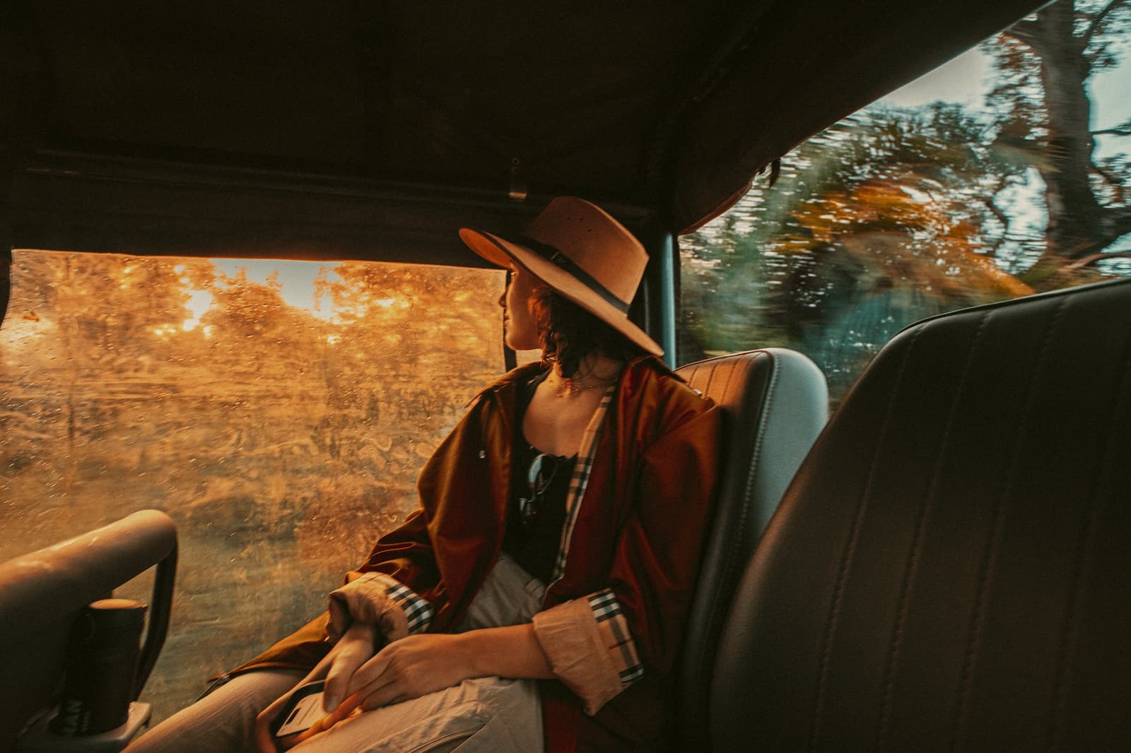 Regen am Fenster, Gold auf allem anderen, die Fahrt ist schon das Abenteuer. Photo: Felipe Castellari Eine Reisende mit breiter Krempe und rostfarbenem Poncho blickt durch ein regengetupftes Fenster eines 4x4-Safarifahrzeugs, goldenes Licht füllt den Innenraum während einer Luxus-Pantanal-und-Amazonas-Reise