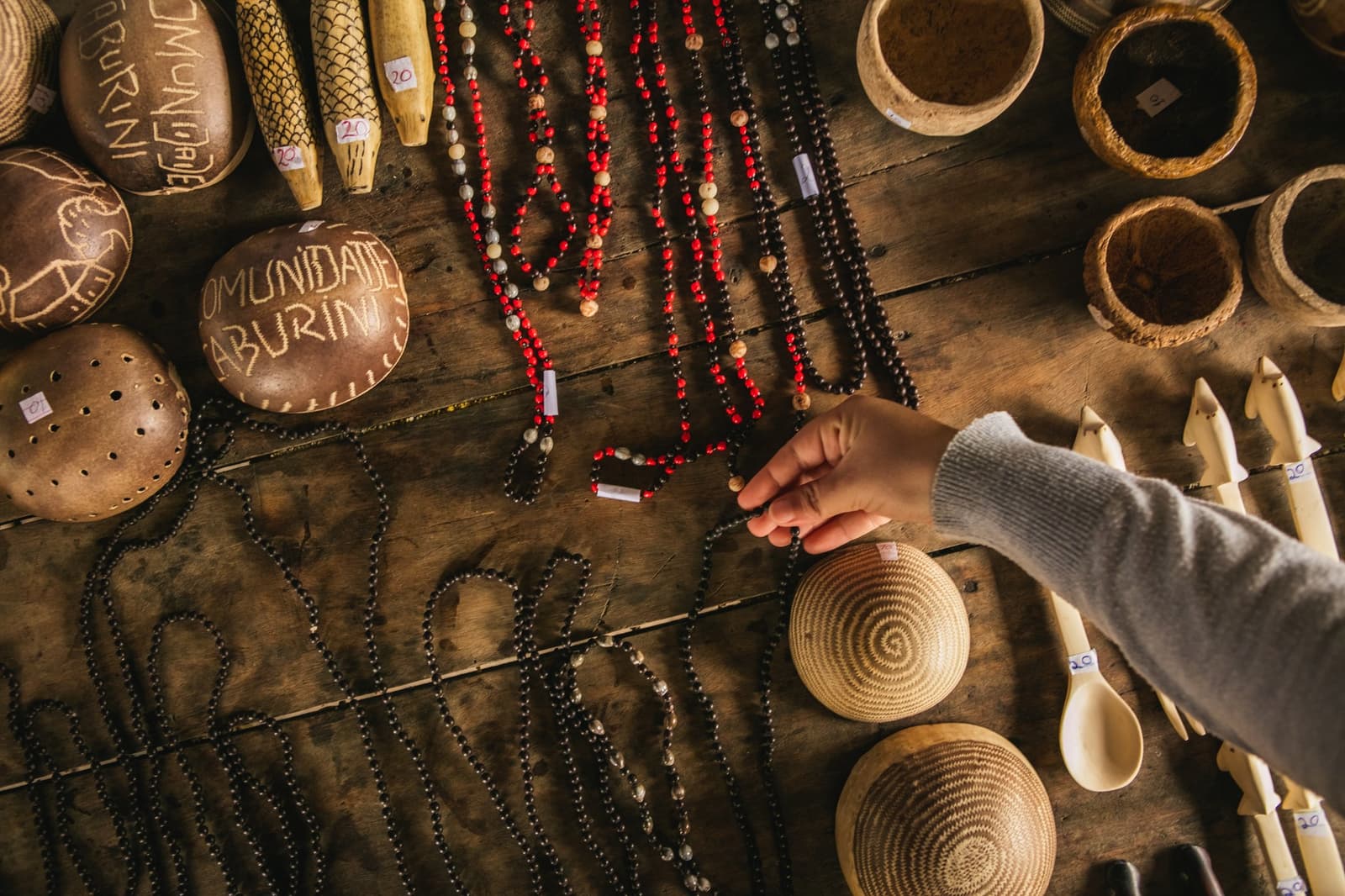 Alles op tafel heeft een maker met een naam en een verhaal. Photo: Guilherme Gomes Bovenaanzicht van een houten tafel bedekt met handgemaakt ambachtelijk werk van Comunidade Caburini: rode en donkere zaadbadkraalnecklaces, gesneden kalebassen, gevlochten palmmanden, houten lepels en beenderen figuurtjes, terwijl een hand van een bezoeker ernaar reikt