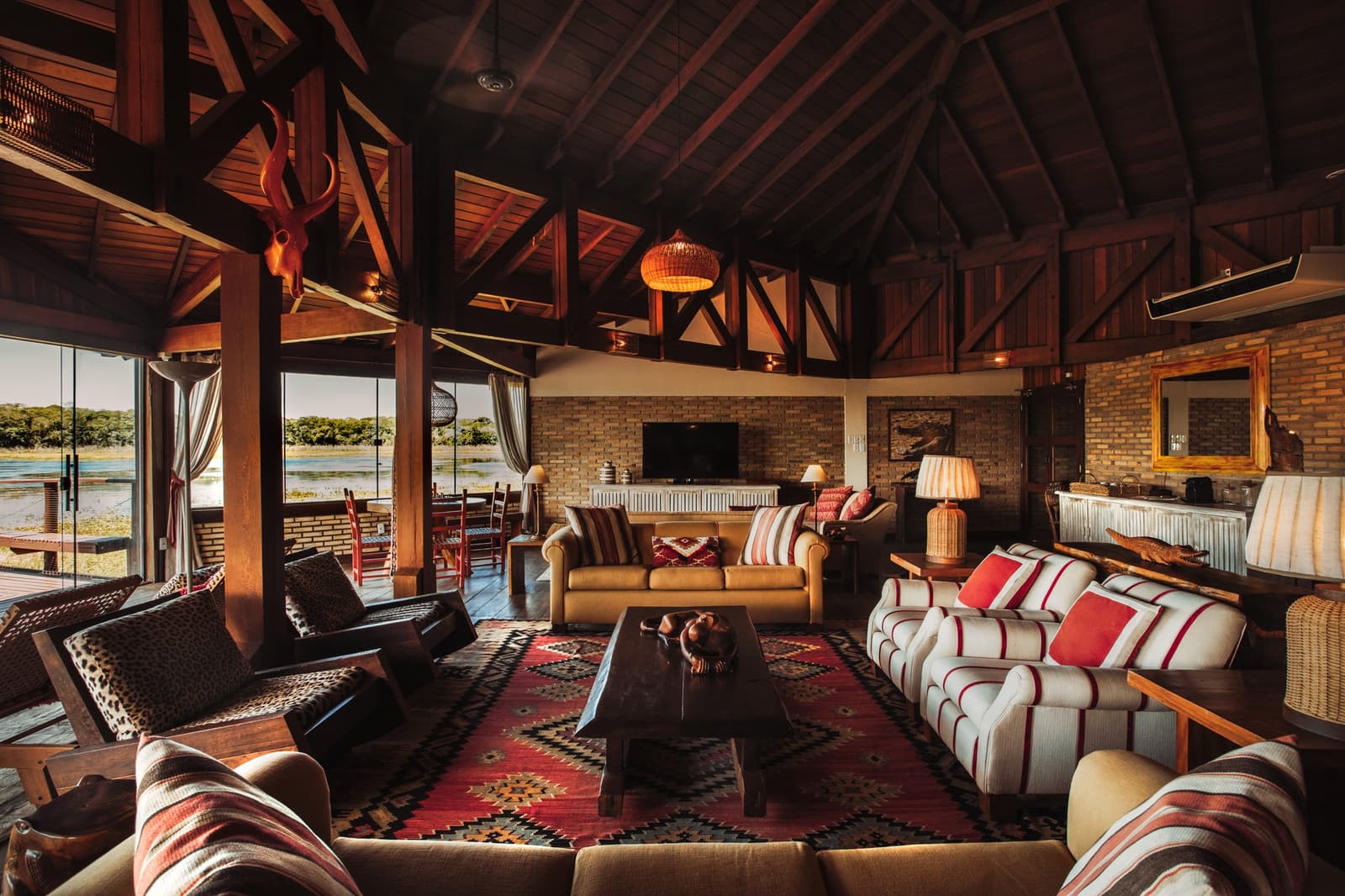 Ankommen, einsinken, den Fluss durch die offenen Türen beobachten, so fängt ein guter Abend an. Photo: Felipe Castellari Geräumiger Aufenthaltsraum einer Lodge auf einer Luxury Pantanal and Amazon Trip mit freiliegenden dunklen Holzdachbalken, gewebter Hängeleuchte, Kelim-Teppich, Leder- und Streifensofas, Tierschädel-Wanddekoration und offenen Glastüren mit Blick auf den Fluss