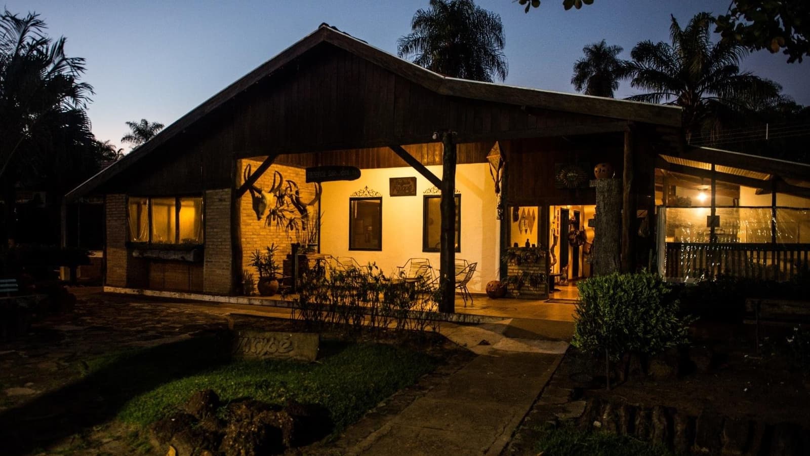 Het avondeten staat bijna klaar, daarna is de nacht aan de beesten. Een rustieke houten lodge die bij schemering warm barnsteenkleurig licht uitstraalt, met een open veranda, wanddecoraties en palmbomen als silhouet tegen een diepblauwe avondlucht