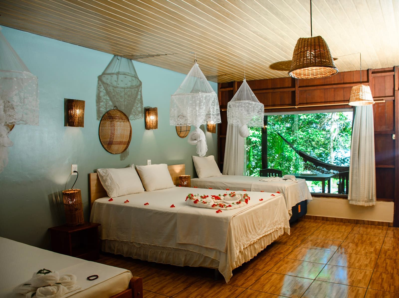 Bloemblaadjes op het bed, een hangmat buiten het raam. De jungle kan even wachten. Comfortabele kamer in een jungle lodge op een Amazon- en Pantanaltour, met twee witte bedden versierd met rode bloemblaadjes en handdoekkunst, kanten klamboes aan het plafond, rieten hanglampen en een groot raam met uitzicht op een hangmat omringd door groen bos